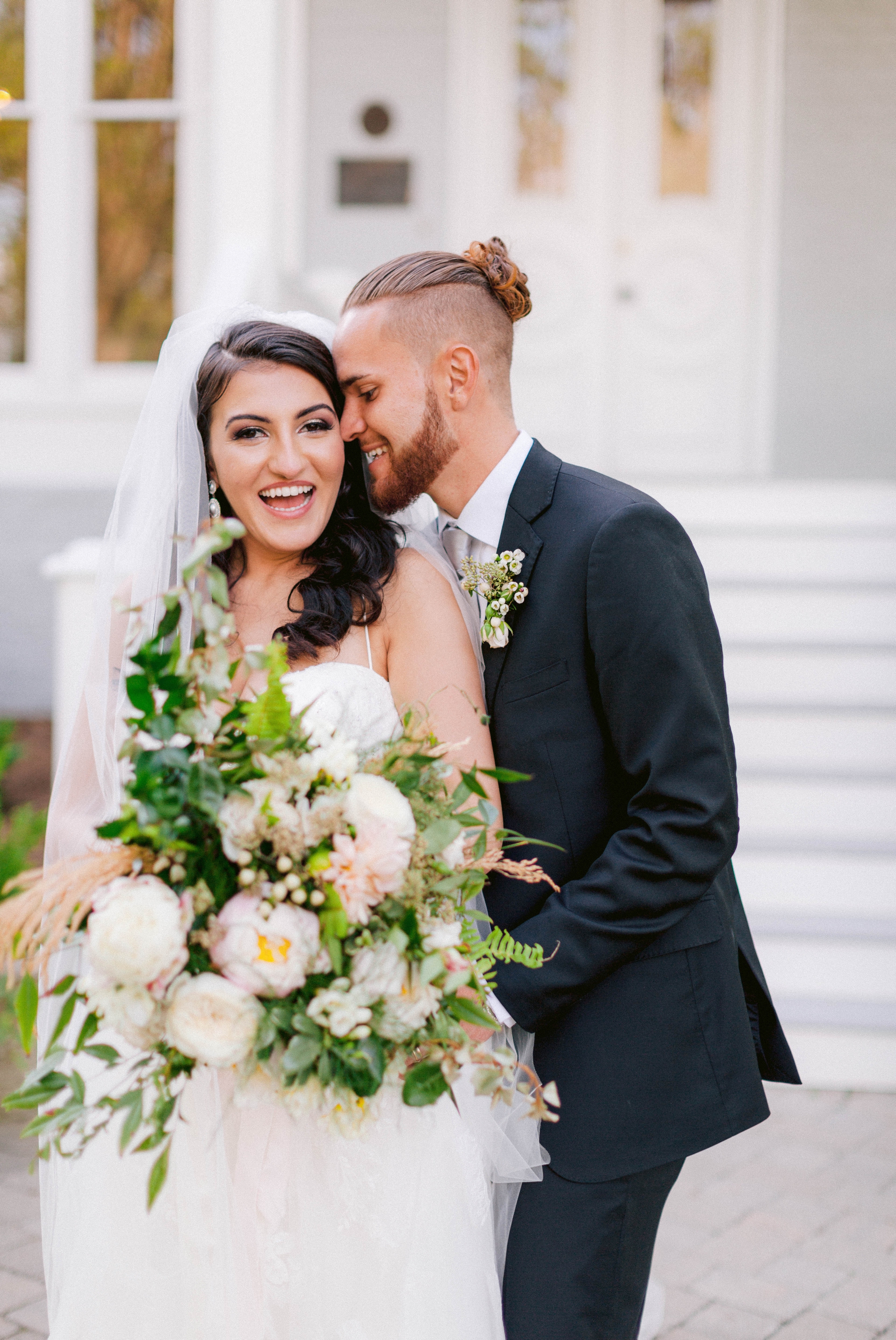  Bride and groom laughing - Wedding Portraits on the front porch of an all white luxury estate - Bride is wearing a Aline Ballgown by Cherish by Southern Bride with a long cathedral veil - Groom is wearing a black suit by Generation Tux and has a man