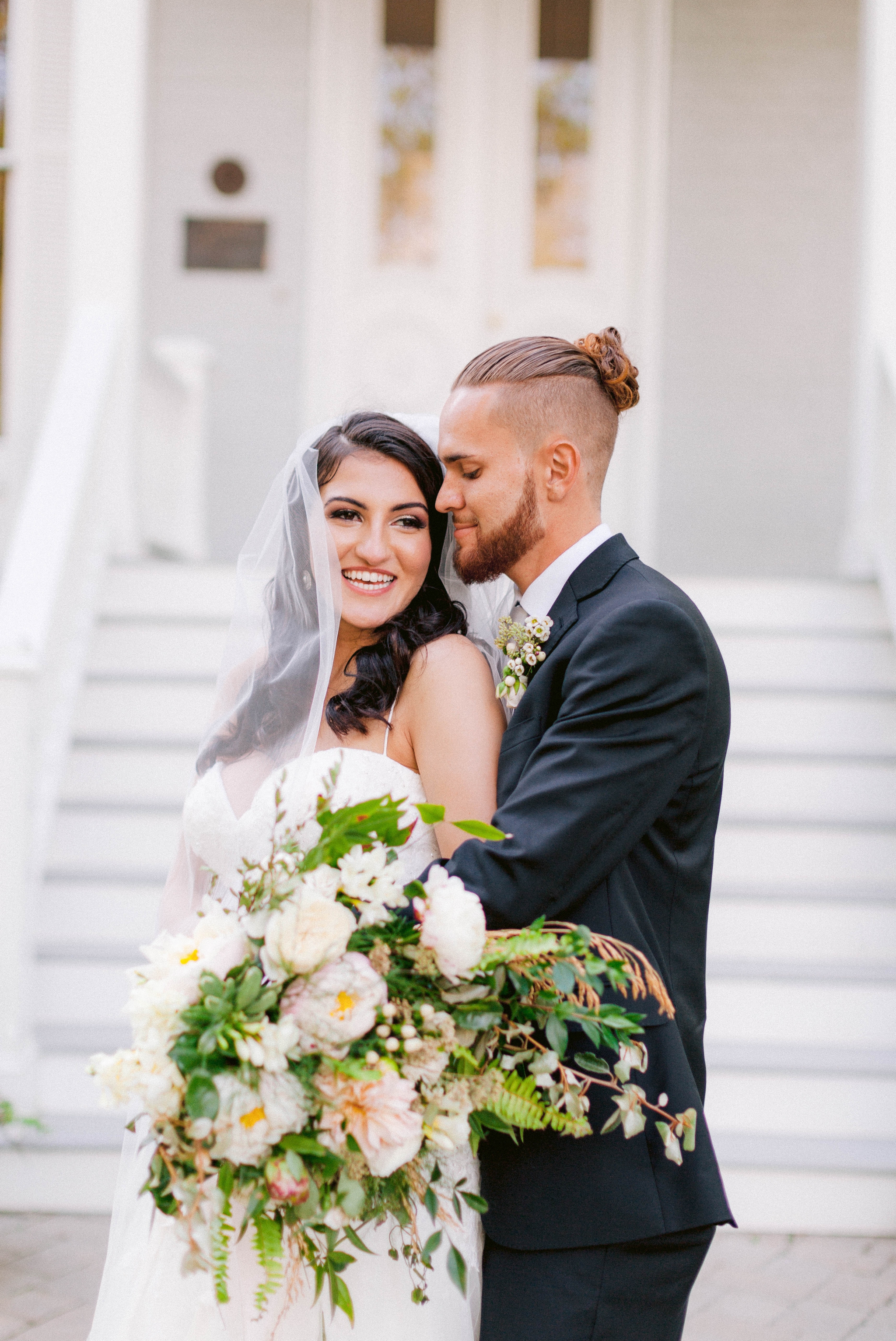  Bride and groom laughing - Wedding Portraits on the front porch of an all white luxury estate - Bride is wearing a Aline Ballgown by Cherish by Southern Bride with a long cathedral veil - Groom is wearing a black suit by Generation Tux and has a man