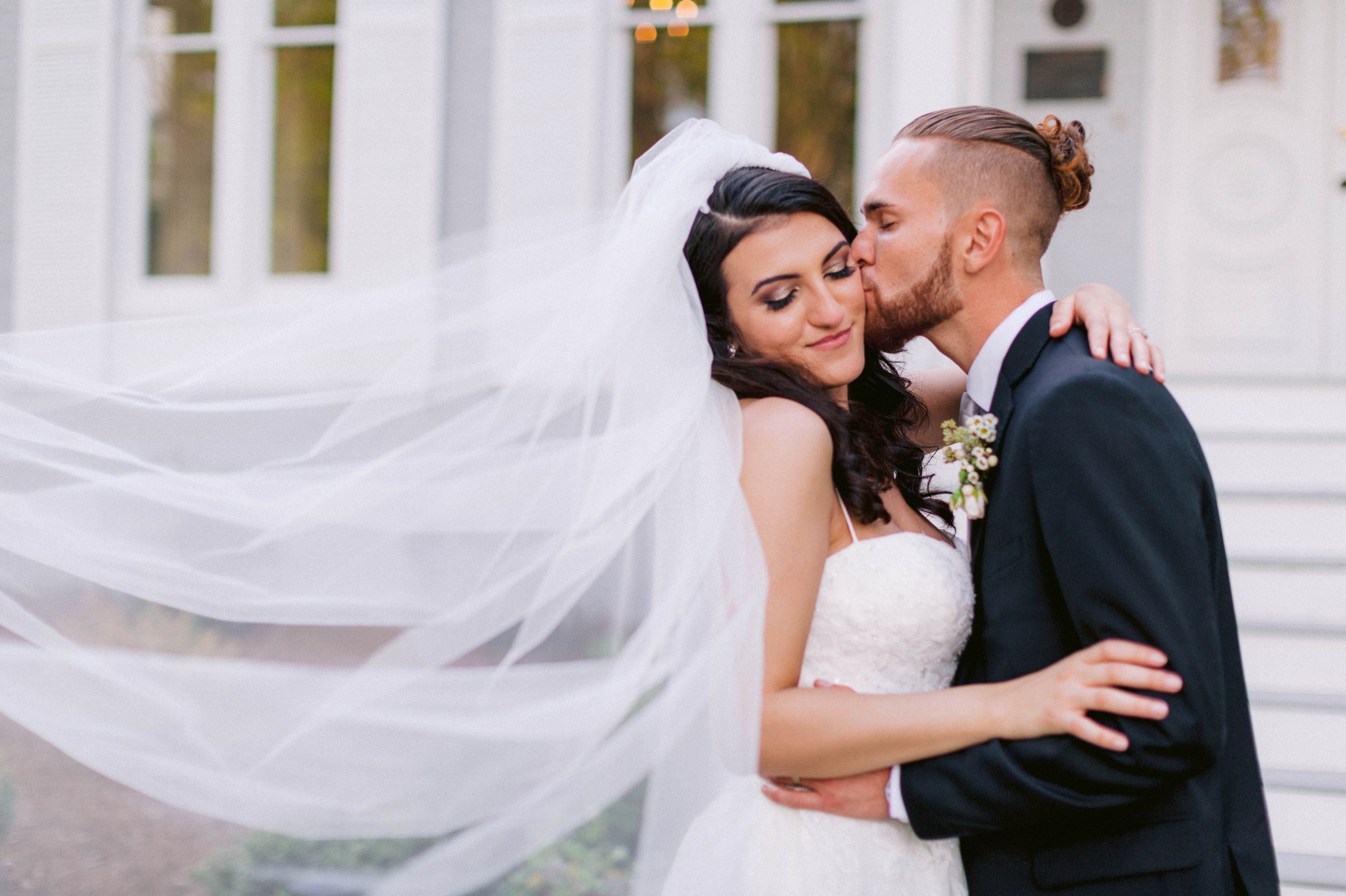  Veil shot - Wedding Portraits on the front porch of an all white luxury estate - Bride is wearing a Aline Ballgown by Cherish by Southern Bride with a long cathedral veil - Groom is wearing a black suit by Generation Tux and has a man bun - Fine Art