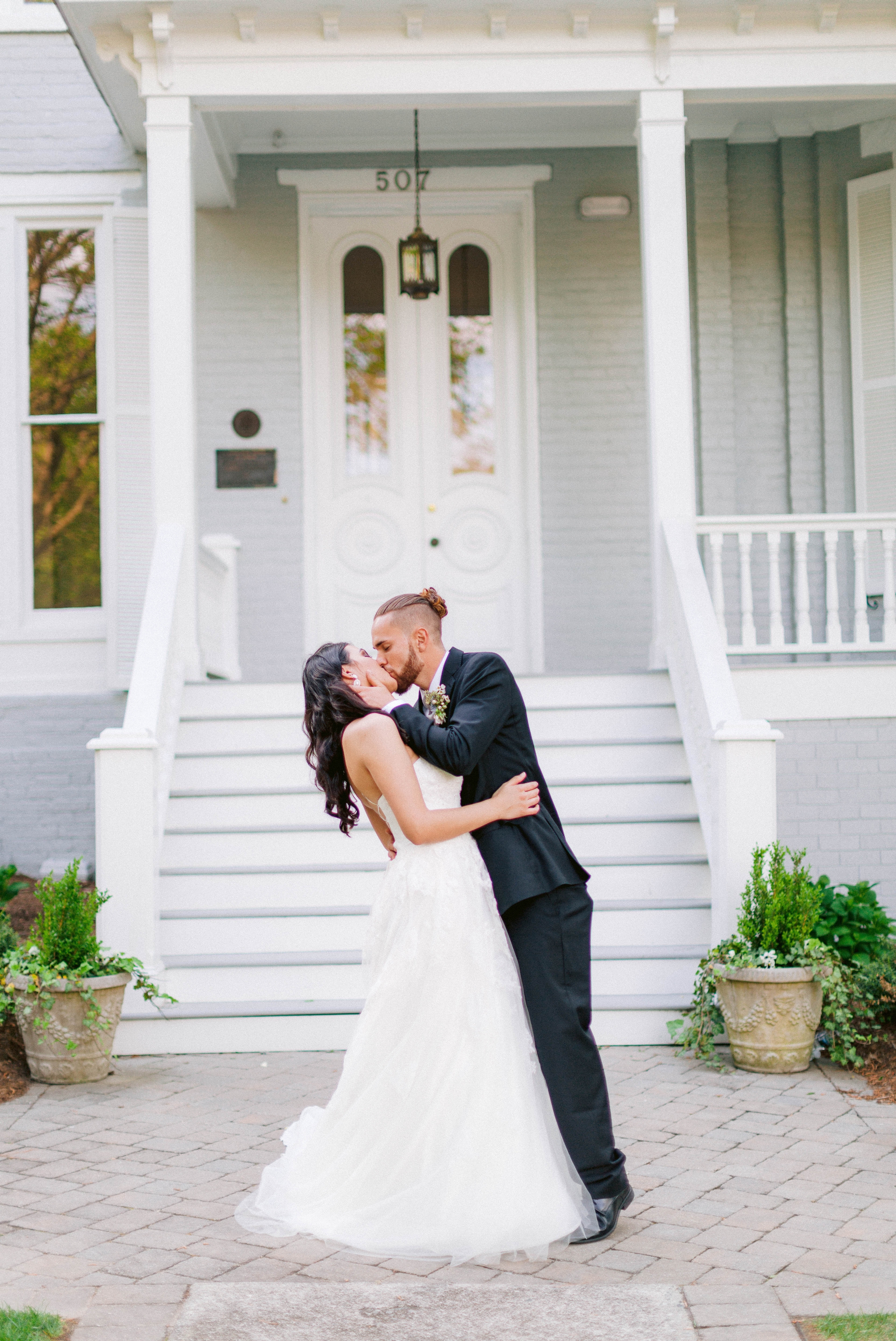  Bride kissing Groom in front of the mansion - Wedding Portraits on the front porch of an all white luxury estate - Bride is wearing a Aline Ballgown by Cherish by Southern Bride with a long cathedral veil - Groom is wearing a black suit by Generatio