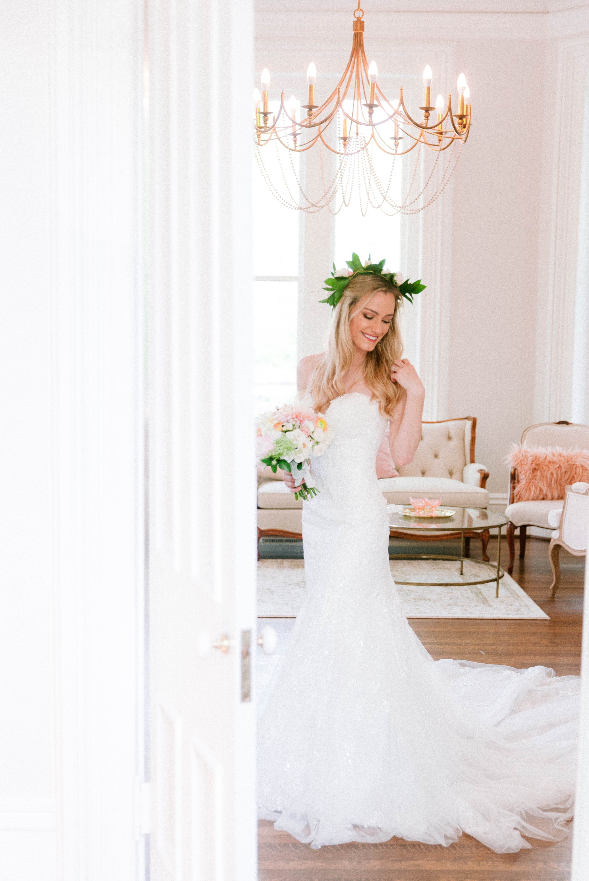 Indoor Bridal Portraits in an all white room at a luxury estate with natural light before the ceremony - Bride is wearing a Hawaiian Flower Crown in a Wedding Gown by Stella York and standing in the doorway with a golden chandelier with vintage furn