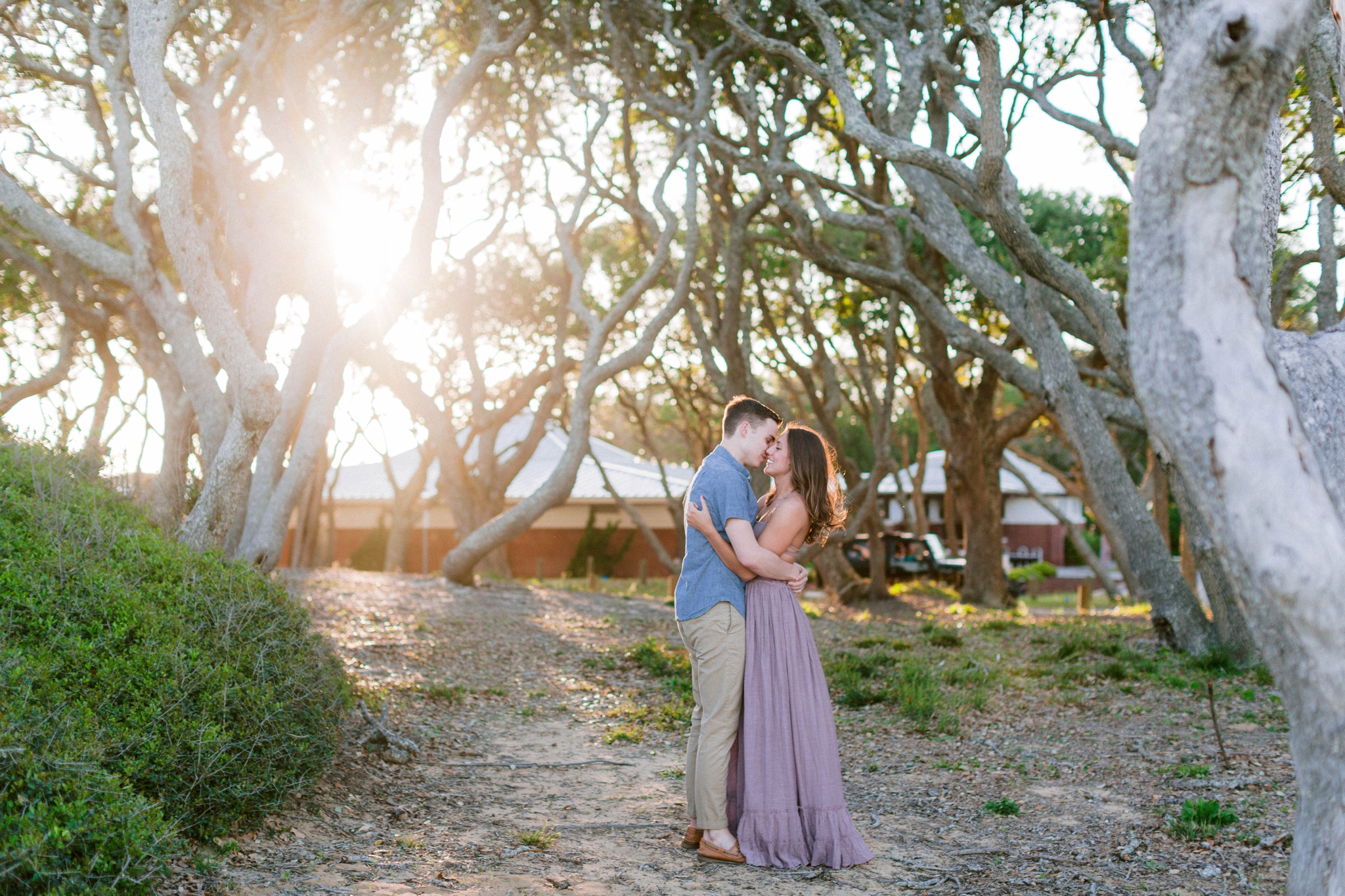  Guy kissing a girl in front of live oak trees with the sunset behind them - Woman is in a flowy pastel maxi dress  - outdoor golden light session - engagement photographer in honolulu, oahu, hawaii - johanna dye photography 