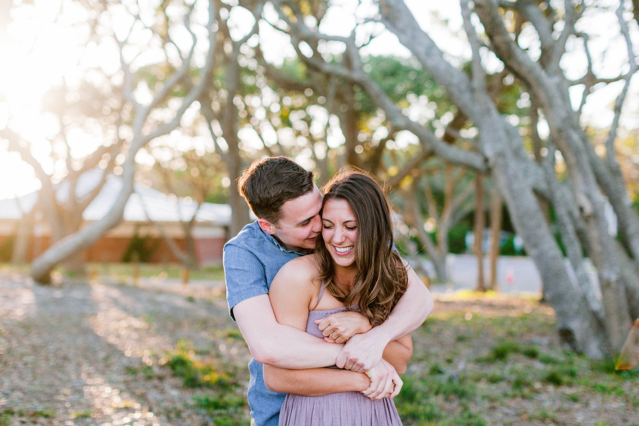  Guy kissing a girl in front of live oak trees with the sunset behind them - Woman is in a flowy pastel maxi dress - outdoor golden light session - engagement photographer in honolulu, oahu, hawaii - johanna dye photography 