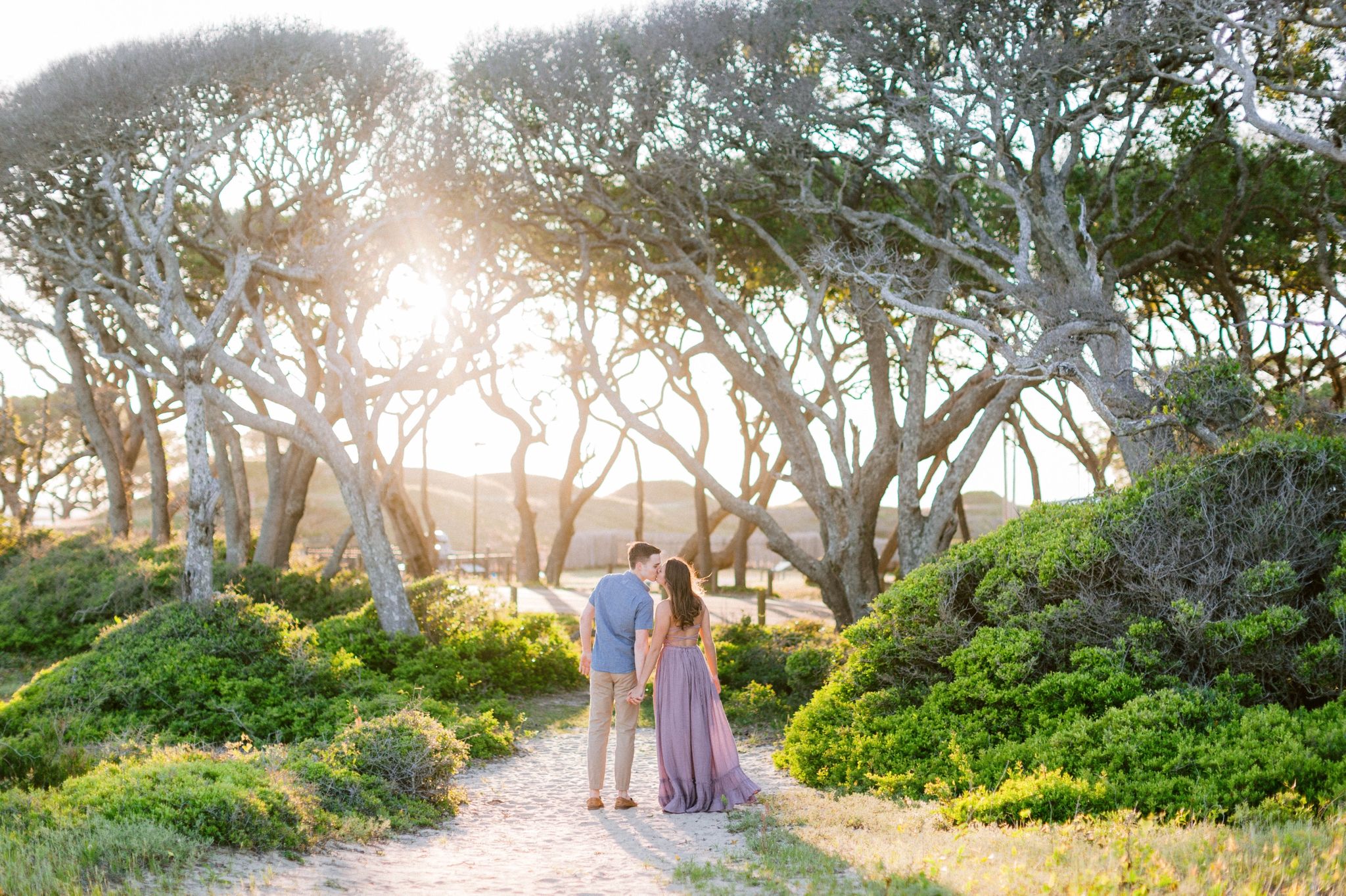  a couple walking towards the sun in front of live oak trees with the sunset behind them - Woman is in a flowy pastel maxi dress - unposed and candid outdoor golden light session - engagement photographer in honolulu, oahu, hawaii - johanna dye 