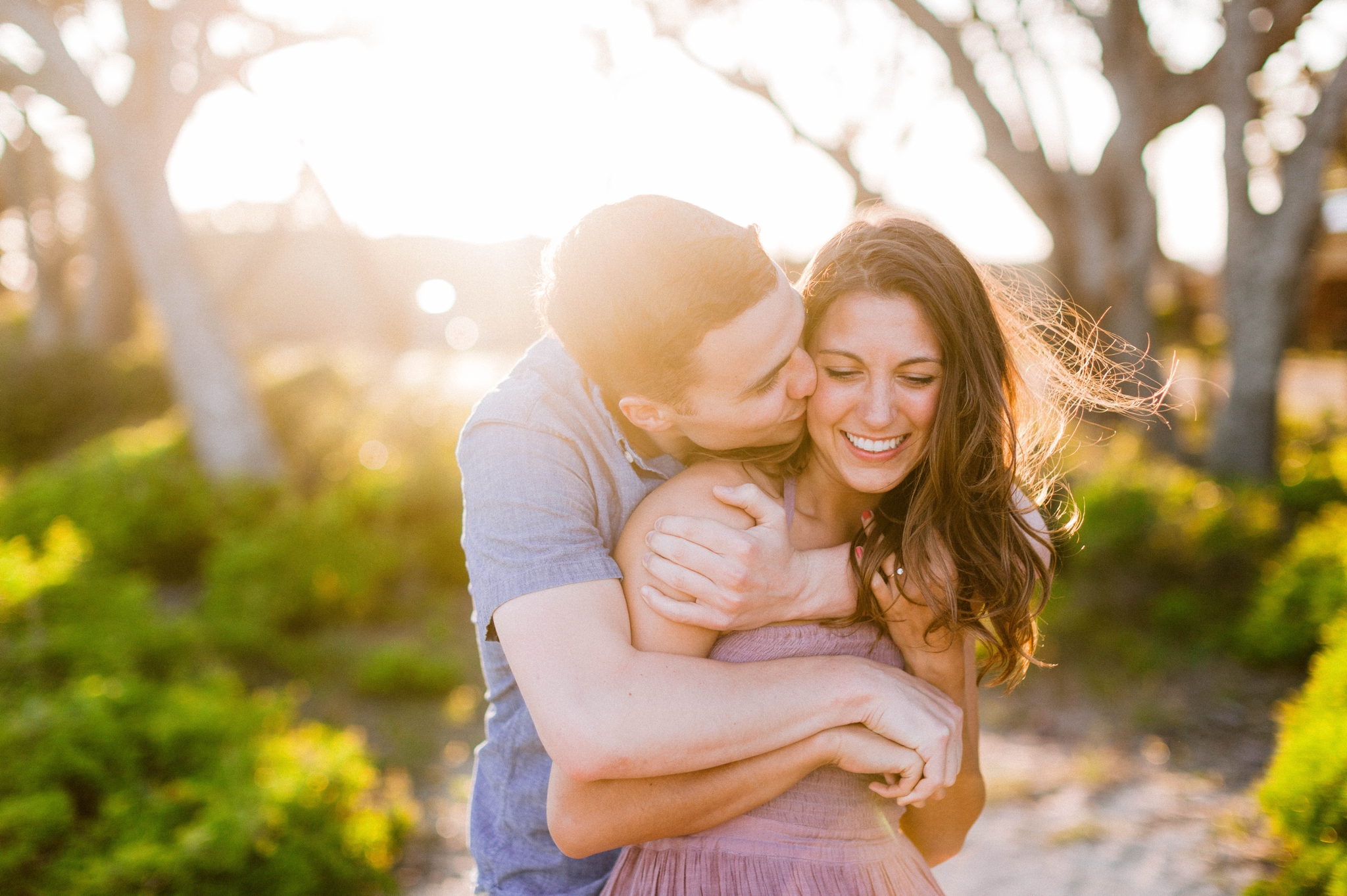  Guy kissing a girl in front of live oak trees with the sunset behind them - Woman is in a flowy pastel maxi dress - outdoor golden light session - engagement photographer in honolulu, oahu, hawaii - johanna dye photography 