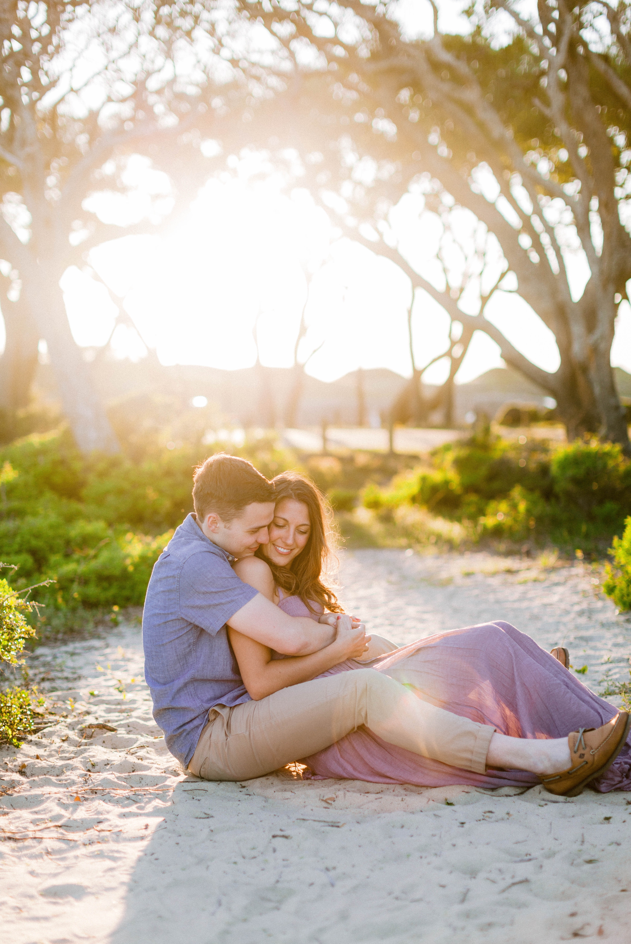  couple giggeling on the beach in front of live oak trees with the sunset behind them - Woman is in a flowy pastel maxi dress - outdoor golden light session - engagement photographer in honolulu, oahu, hawaii - johanna dye photography 