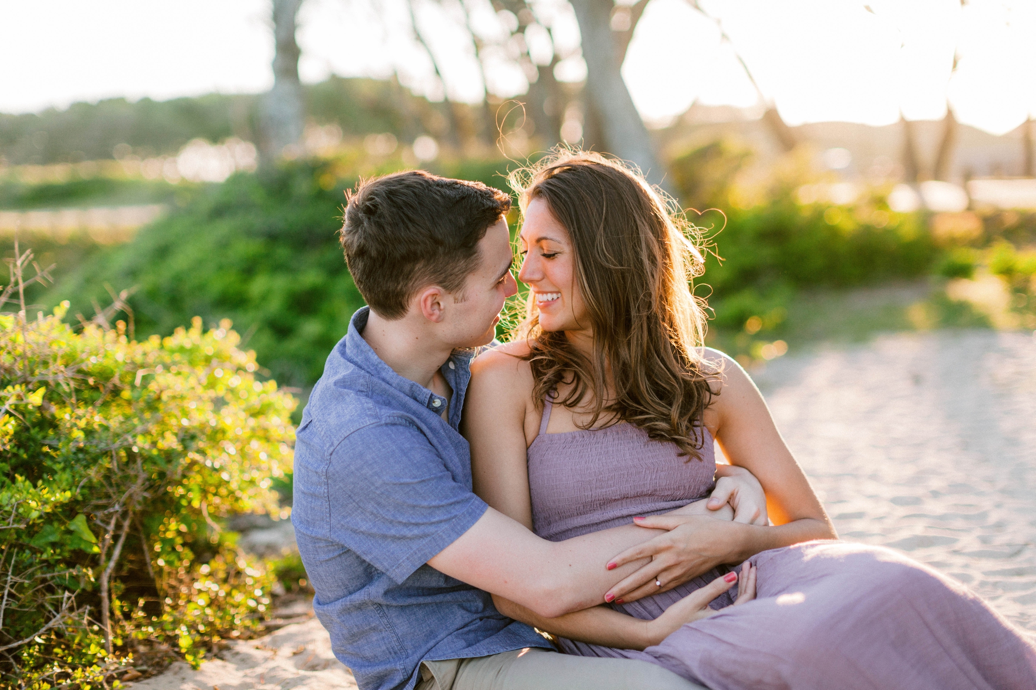  couple cuddeling  on the beach in front of live oak trees with the sunset behind them - Woman is in a flowy pastel maxi dress - outdoor golden light session - engagement photographer in honolulu, oahu, hawaii - johanna dye photography 