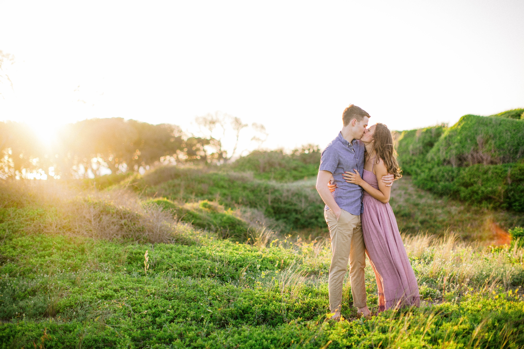  couple kissing in front of lush green dunes and hills with the sunset behind them  - Woman is in a flowy pastel maxi dress - outdoor golden light session - engagement photographer in honolulu, oahu, hawaii - johanna dye photography 