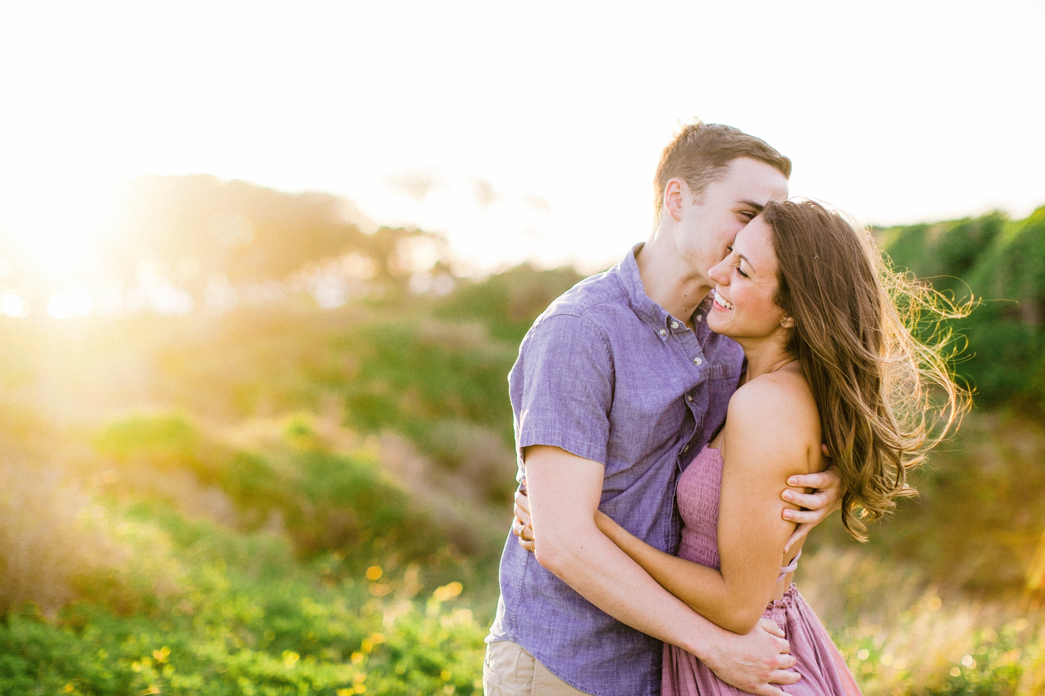  couple kissing in front of lush green dunes and hills with the sunset behind them - Woman is in a flowy pastel maxi dress - outdoor golden light session - engagement photographer in honolulu, oahu, hawaii - johanna dye photography 