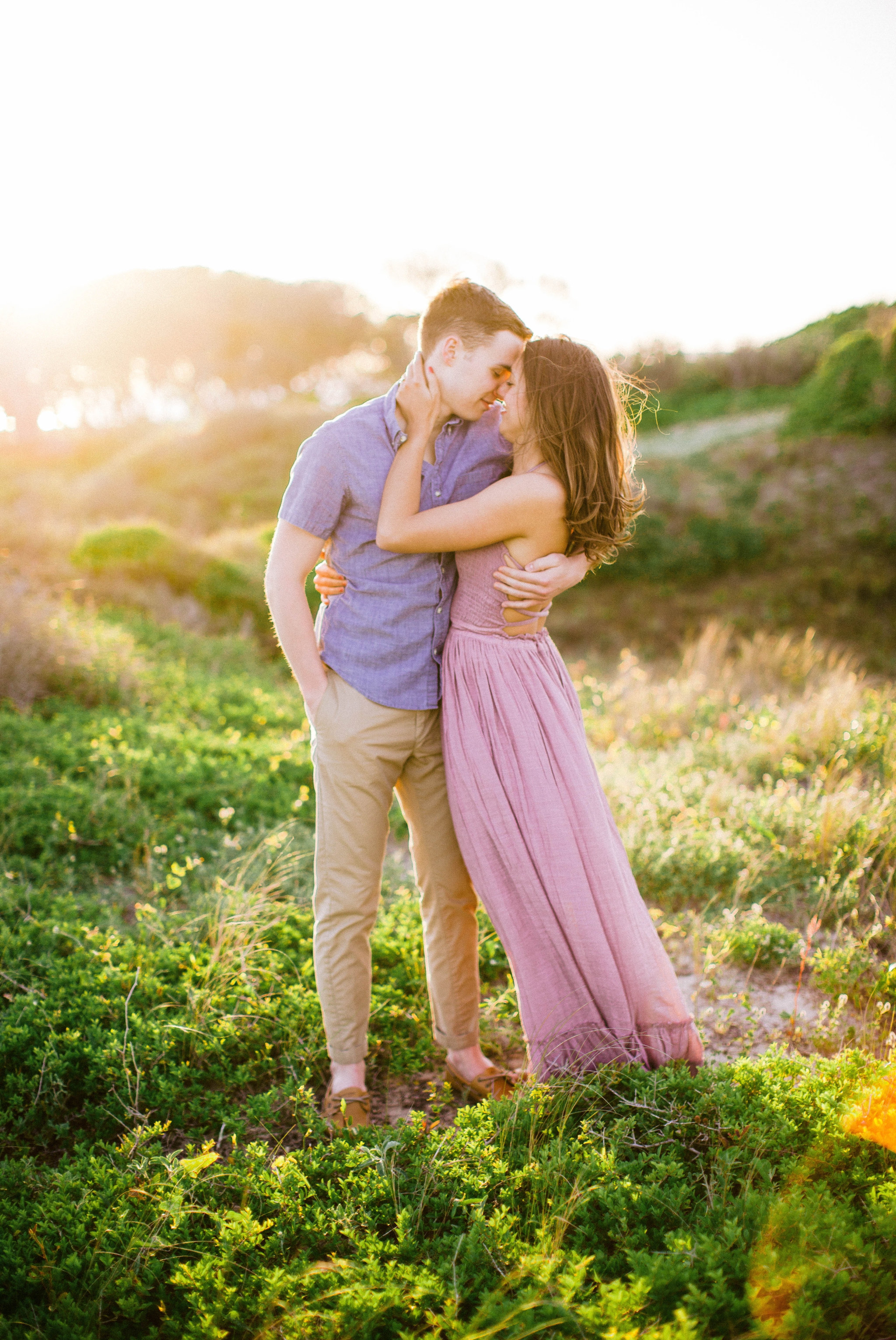  couple kissing in front of lush green dunes and hills with the sunset behind them - Woman is in a flowy pastel maxi dress - outdoor golden light session - engagement photographer in honolulu, oahu, hawaii - johanna dye photography 