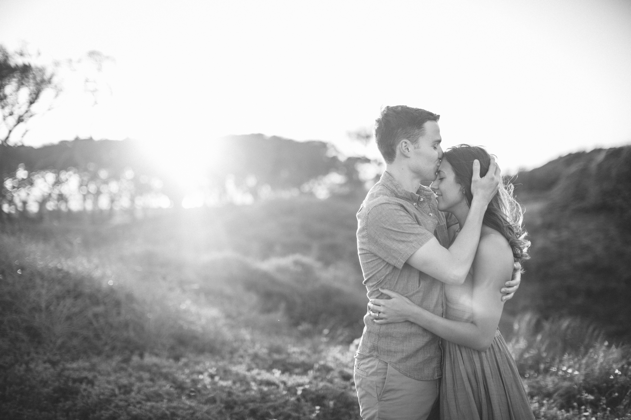  couple looking at each other while his hand is in her hair wile he is kissing her on the forehead- in front of lush green dunes and hills with the sunset behind them - Woman is in a flowy pastel maxi dress - outdoor golden light session - engagement