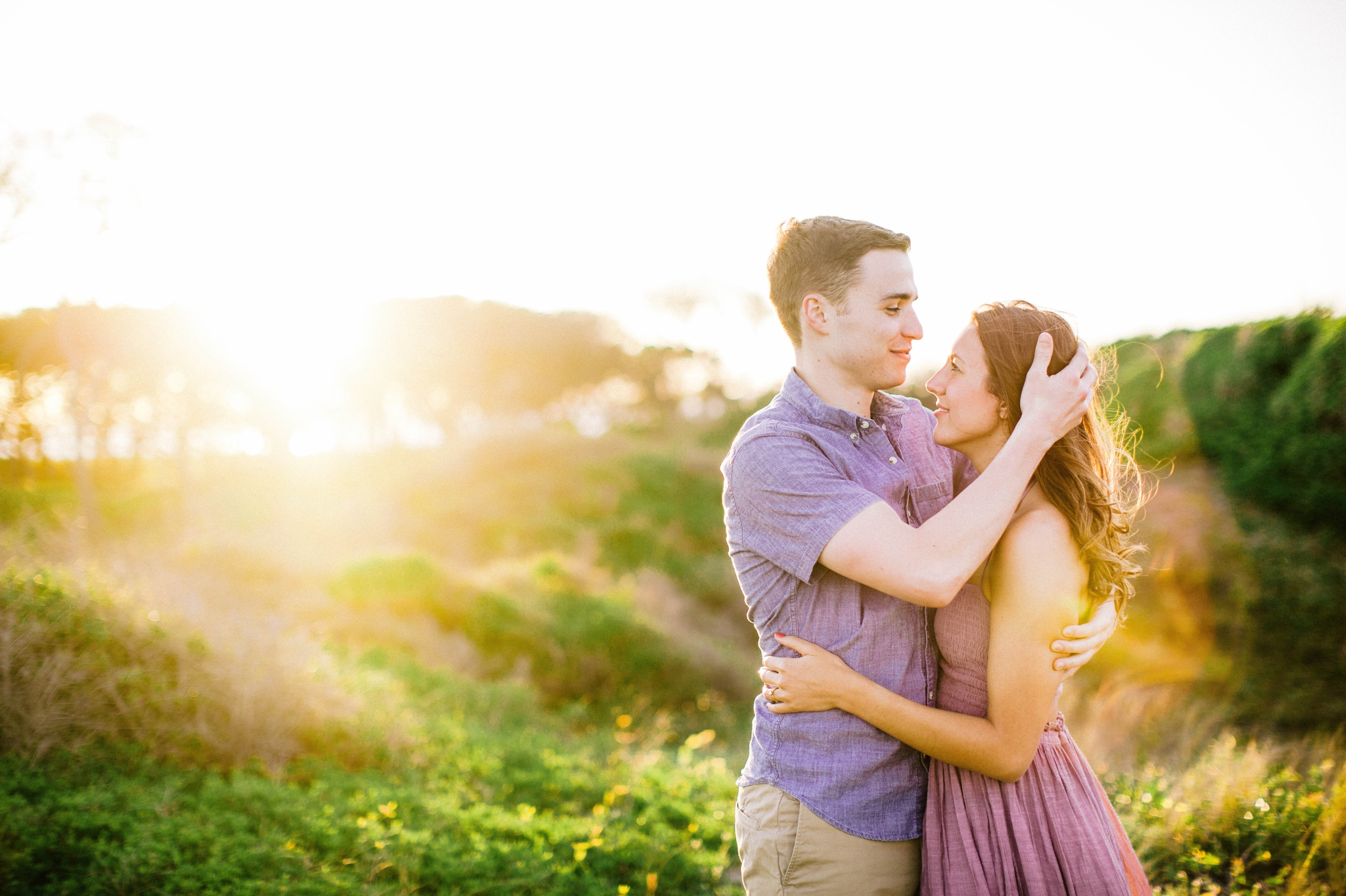  couple looking at each other while his hand is in her hair - in front of lush green dunes and hills with the sunset behind them - Woman is in a flowy pastel maxi dress - outdoor golden light session - engagement photographer in honolulu, oahu, hawai