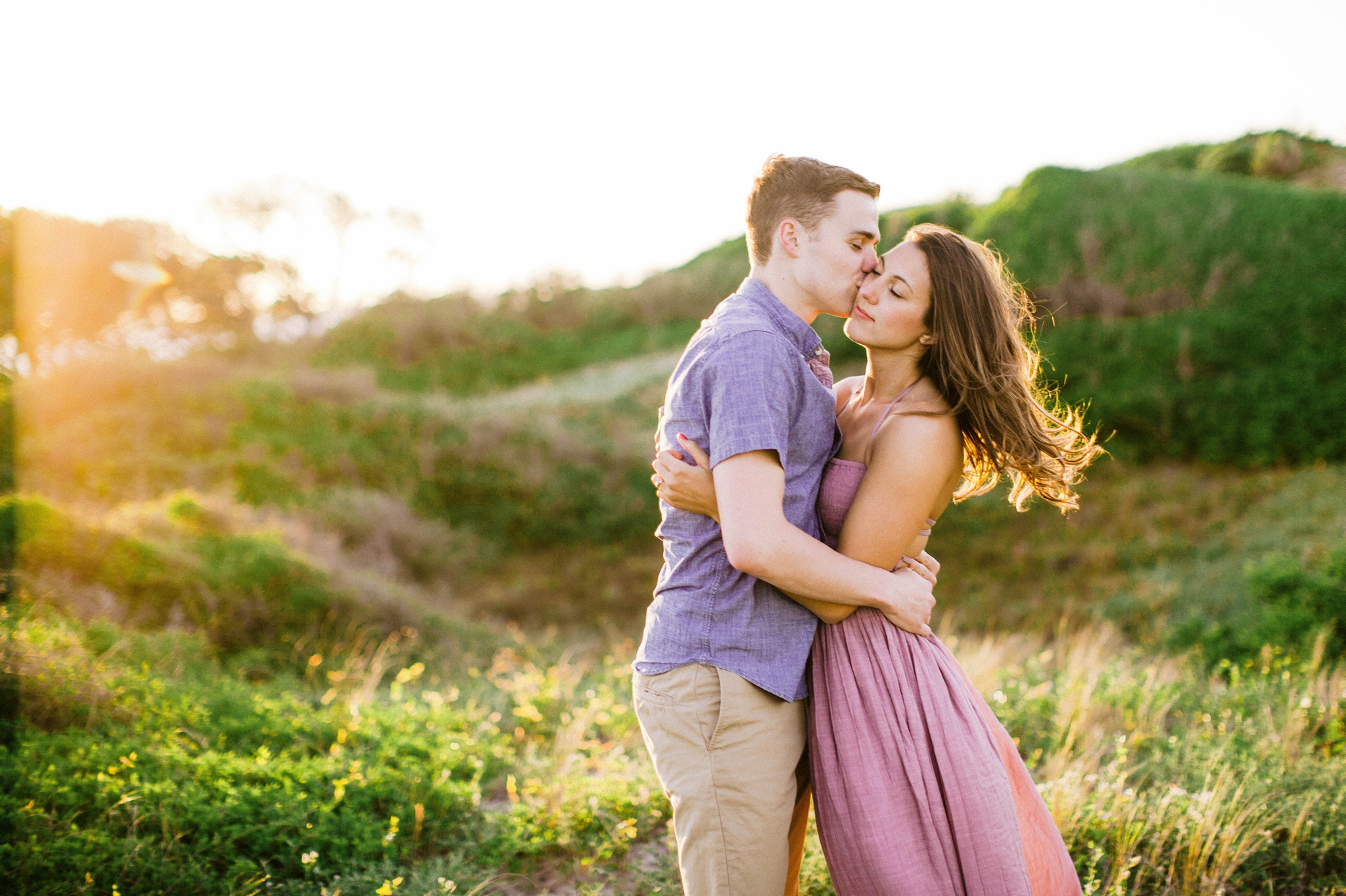 couple looking at each other while his hand is in her hair - in front of lush green dunes and hills with the sunset behind them - Woman is in a flowy pastel maxi dress - outdoor golden light session - engagement photographer in honolulu, oahu, hawai