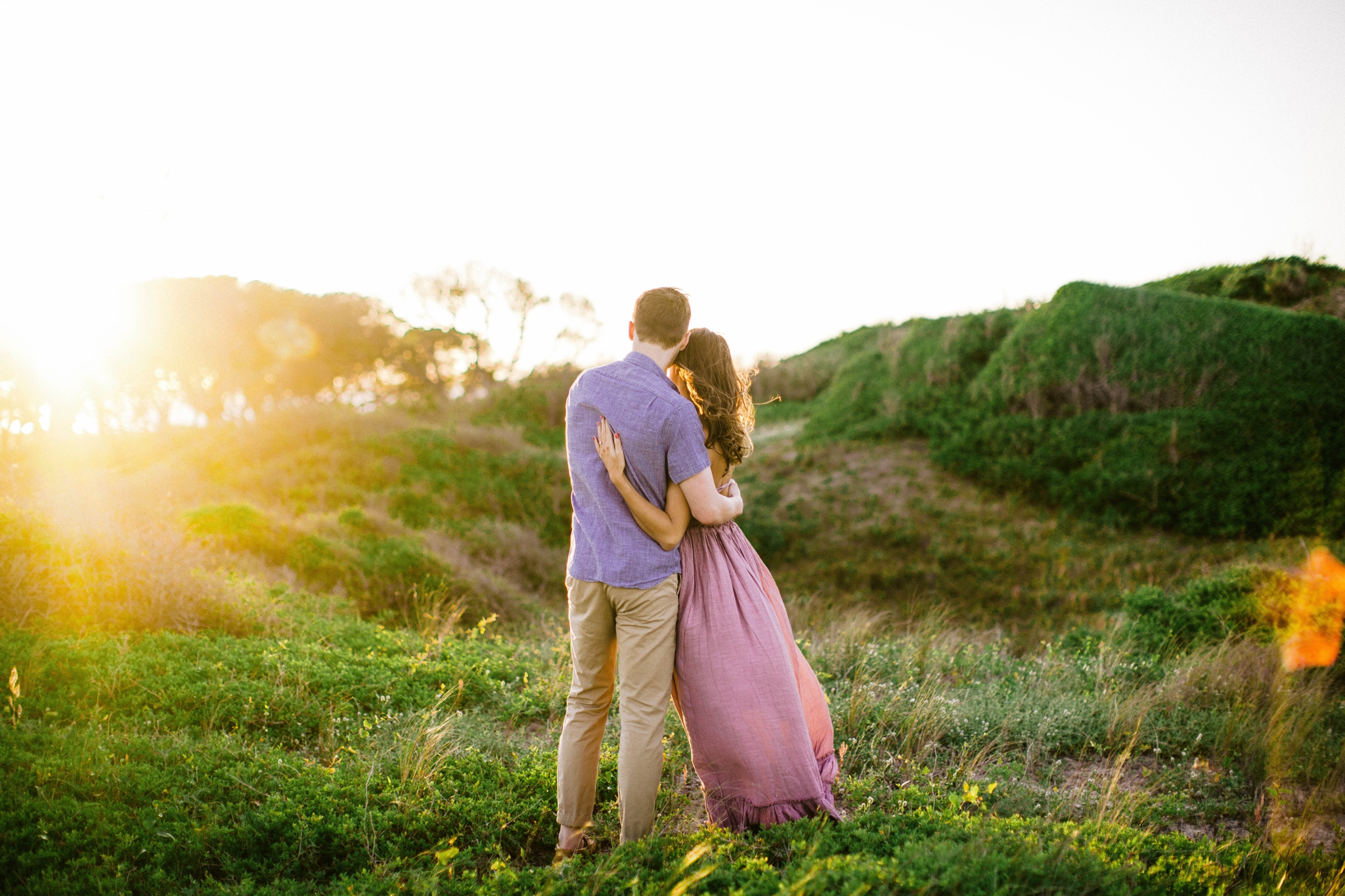  couple looking at the sunset  - in front of lush green dunes and hills with the sunset behind them - Woman is in a flowy pastel maxi dress - candid and unposed outdoor golden light session - engagement photographer in honolulu, oahu, hawaii - johann