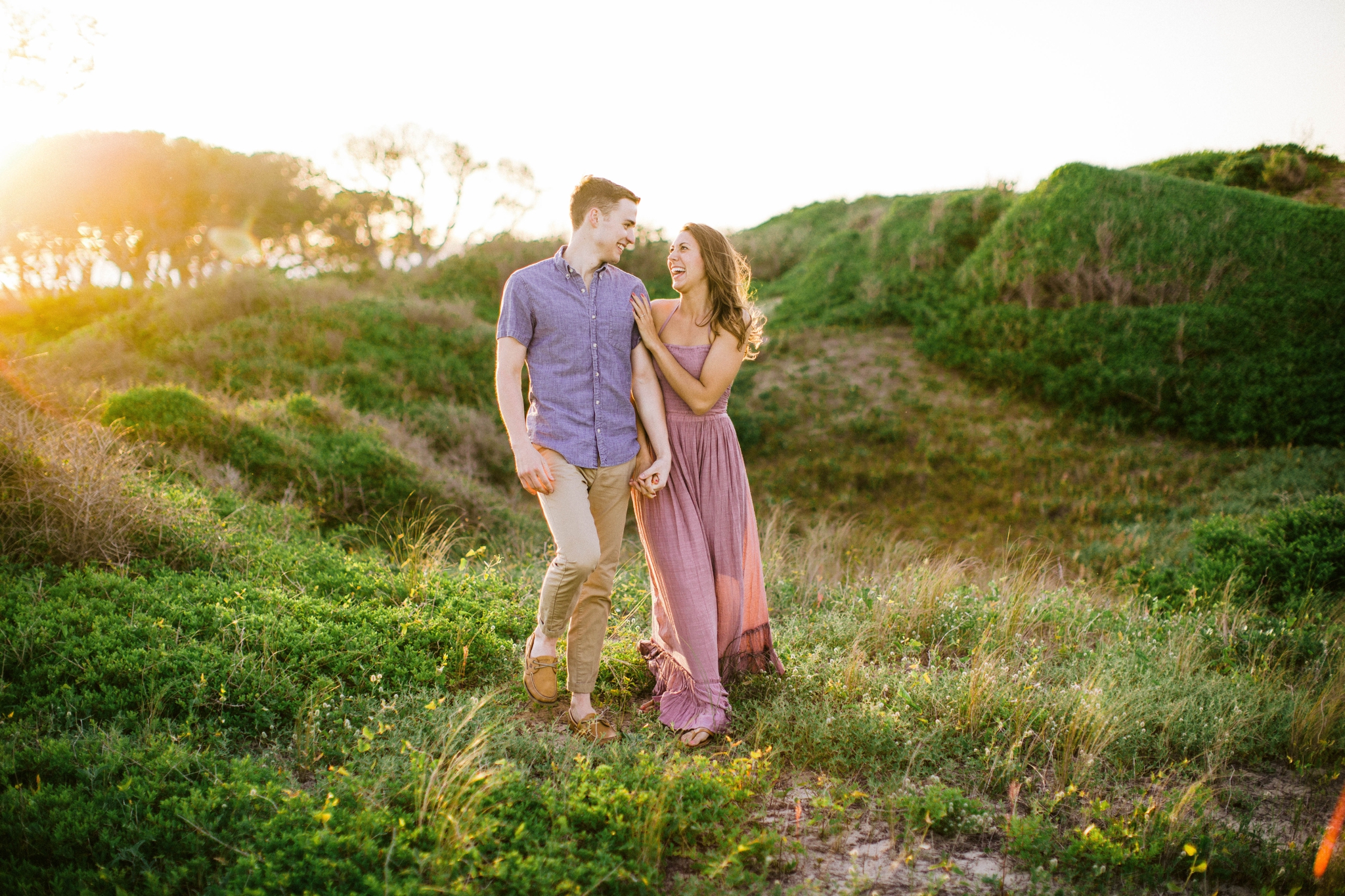  couple walking and laughing - in front of lush green dunes and hills with the sunset behind them - Woman is in a flowy pastel maxi dress - candid and unposed outdoor golden light session - engagement photographer in honolulu, oahu, hawaii - johanna 