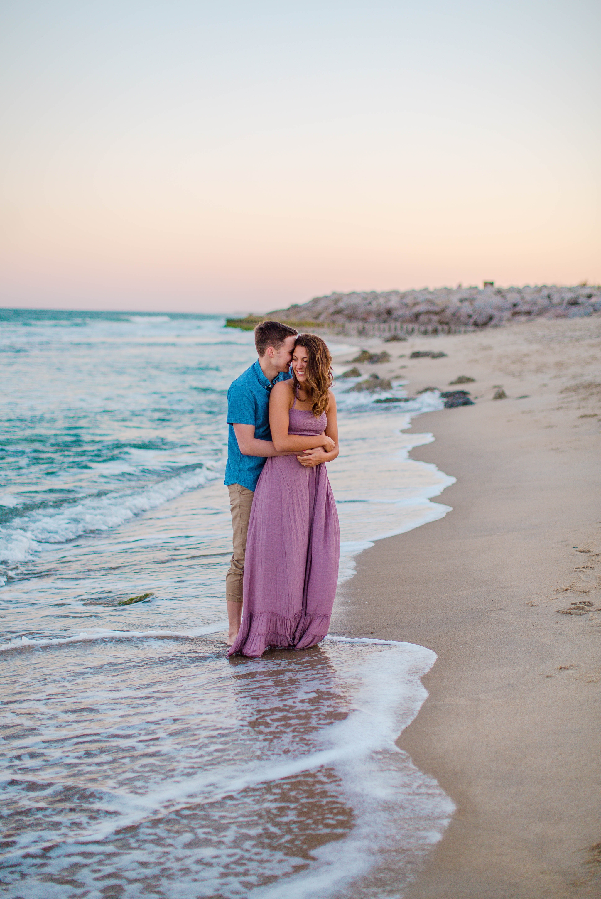  man and woman standing in the water and cuddling - - Woman is in a flowy pastel maxi dress - candid and unposed golden light session - beach engagement photographer in honolulu, oahu, hawaii - johanna dye photography 