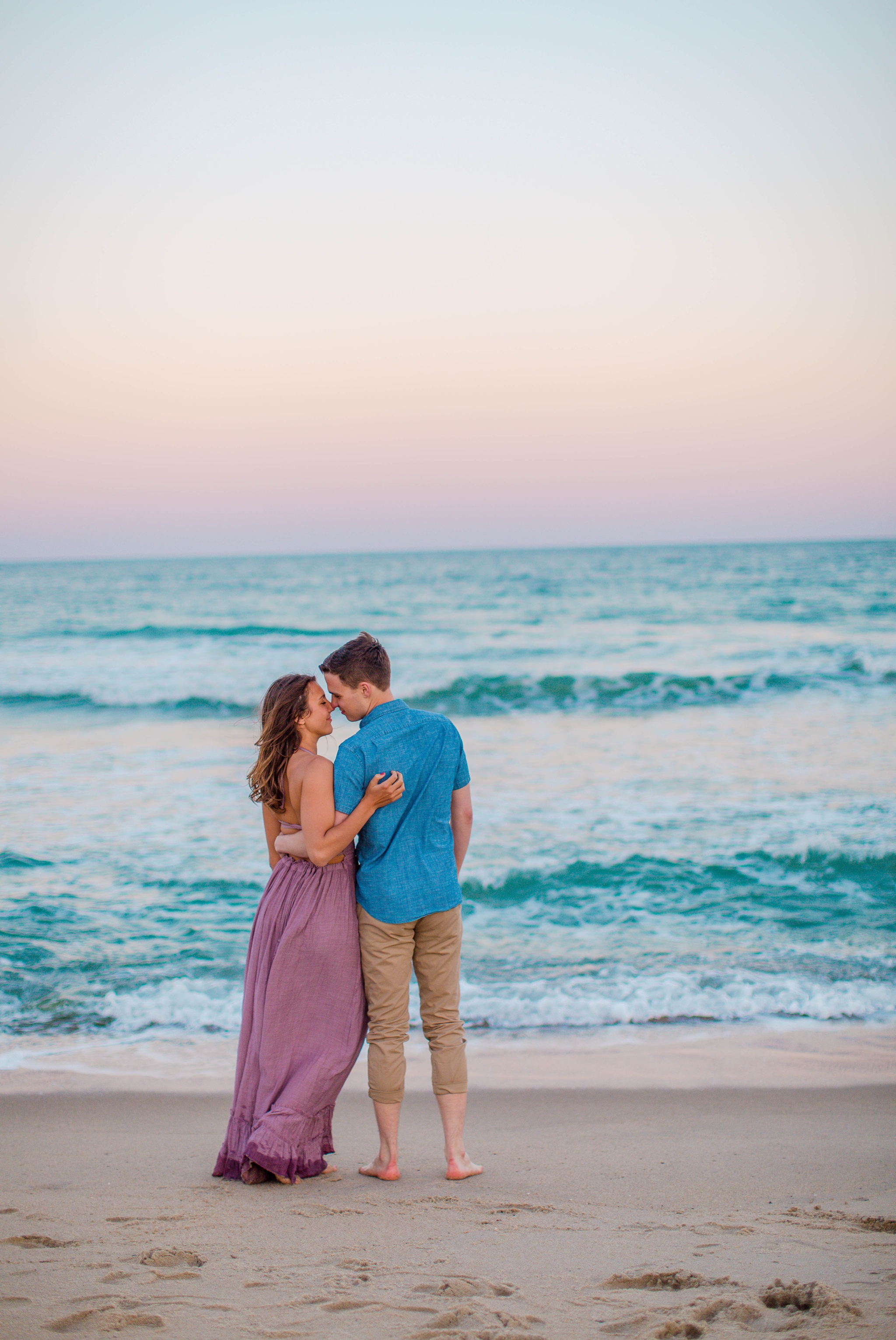  man and woman kissing in front of the sunset - - Woman is in a flowy pastel maxi dress - candid and unposed golden light session - beach engagement photographer in honolulu, oahu, hawaii - johanna dye photography 