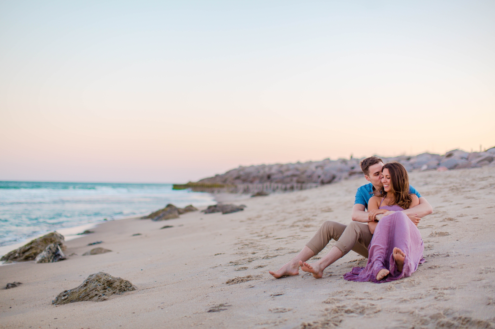  man and woman sitting in the sand - - Woman is in a flowy pastel maxi dress - candid and unposed golden light session - beach engagement photographer in honolulu, oahu, hawaii - johanna dye photography 