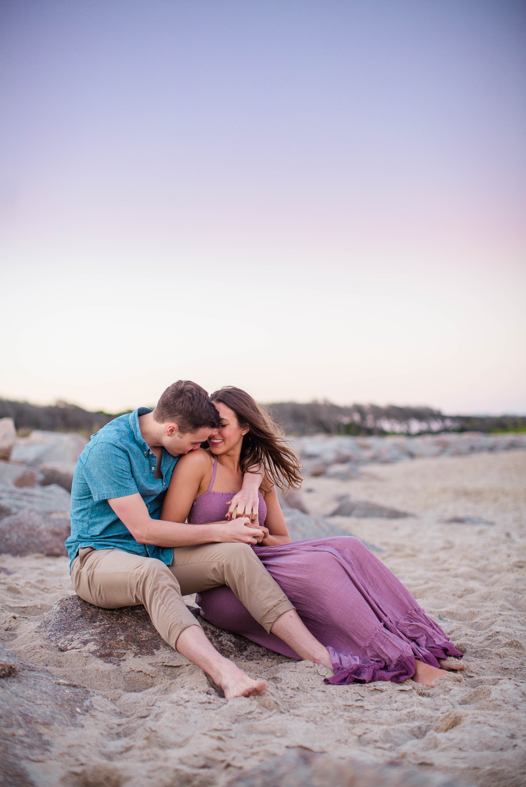  man and woman sitting in the sand kissing - Woman is in a flowy pastel maxi dress - candid and unposed golden light session - beach engagement photographer in honolulu, oahu, hawaii - johanna dye photography 