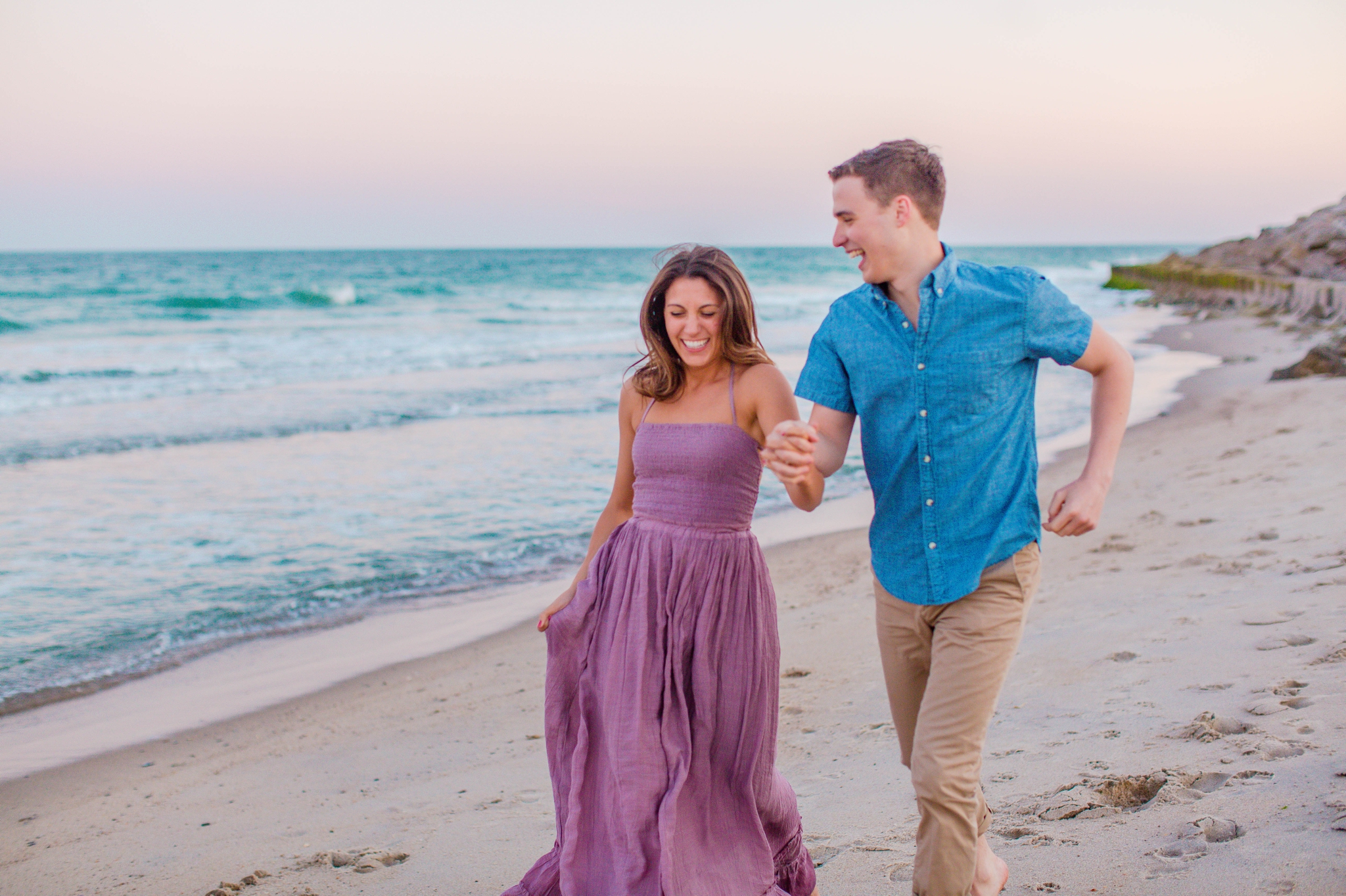  man and woman running on the beach and laughing - Woman is in a flowy pastel maxi dress - candid and unposed golden light session - beach engagement photographer in honolulu, oahu, hawaii - johanna dye photography 