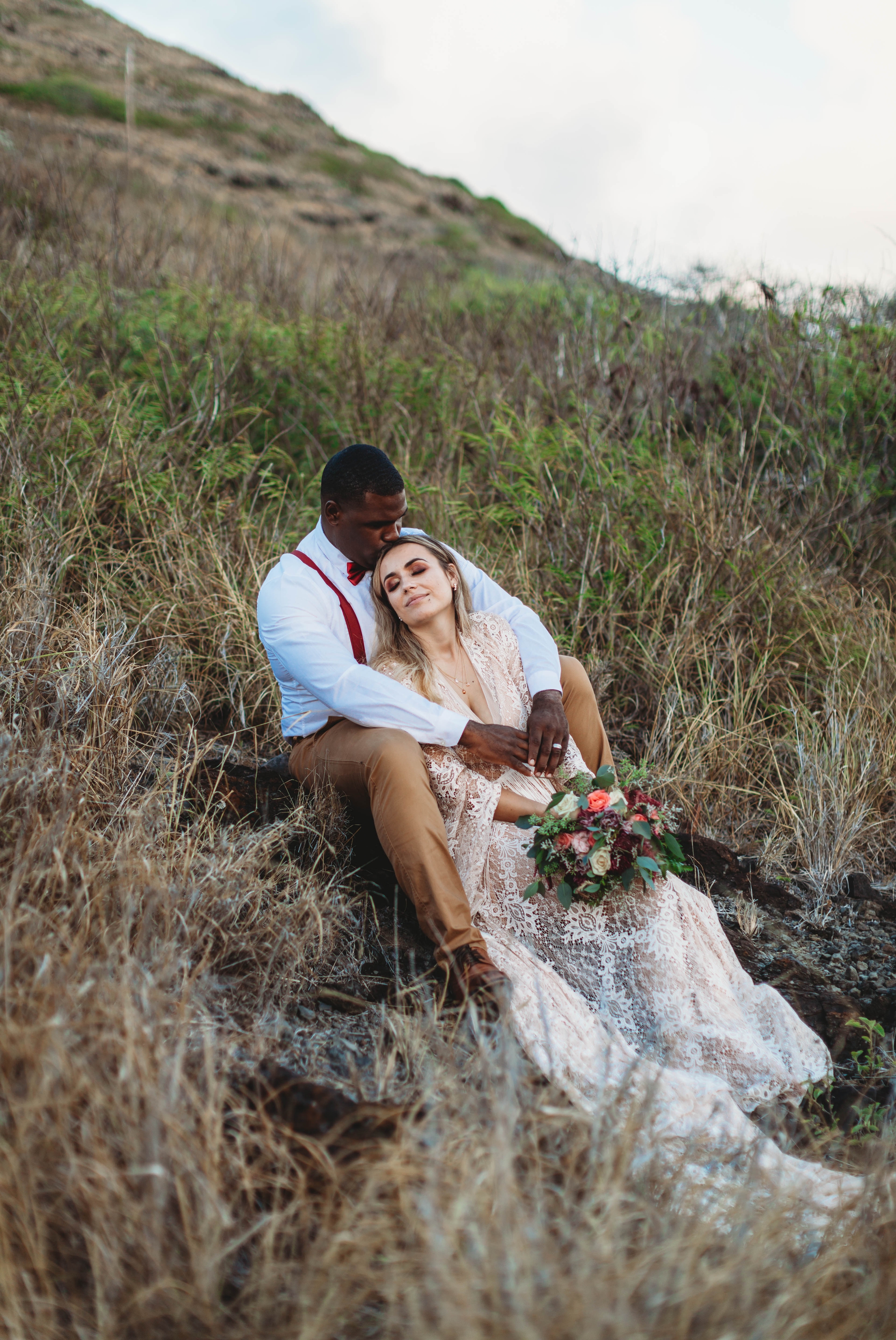  Boho bride and groom sitting in the grass - Bride in a flowy fringe boho wedding dress on the cliffs above - Makapuu Lookout, Waimanalo, HI looking over the ocean with the  Kāohikaipu Island in the background  - Oahu Hawaii Engagement Photographer -