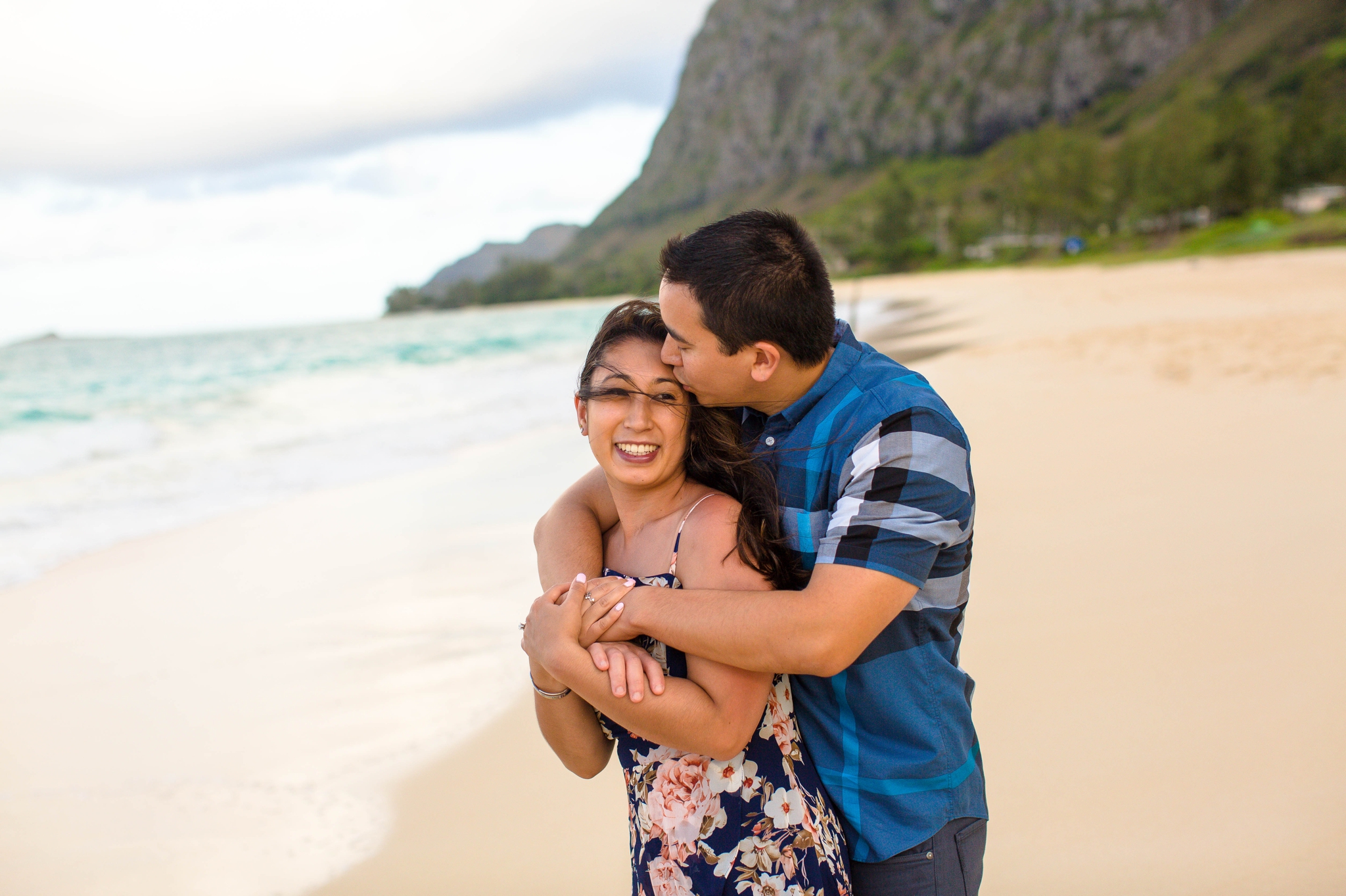 engagement photography session at waimanalo beach park - couple is kissing and snuggeling up to each other in the sand with the ocean and the mountains in the back ground - oahu hawaii wedding photographer