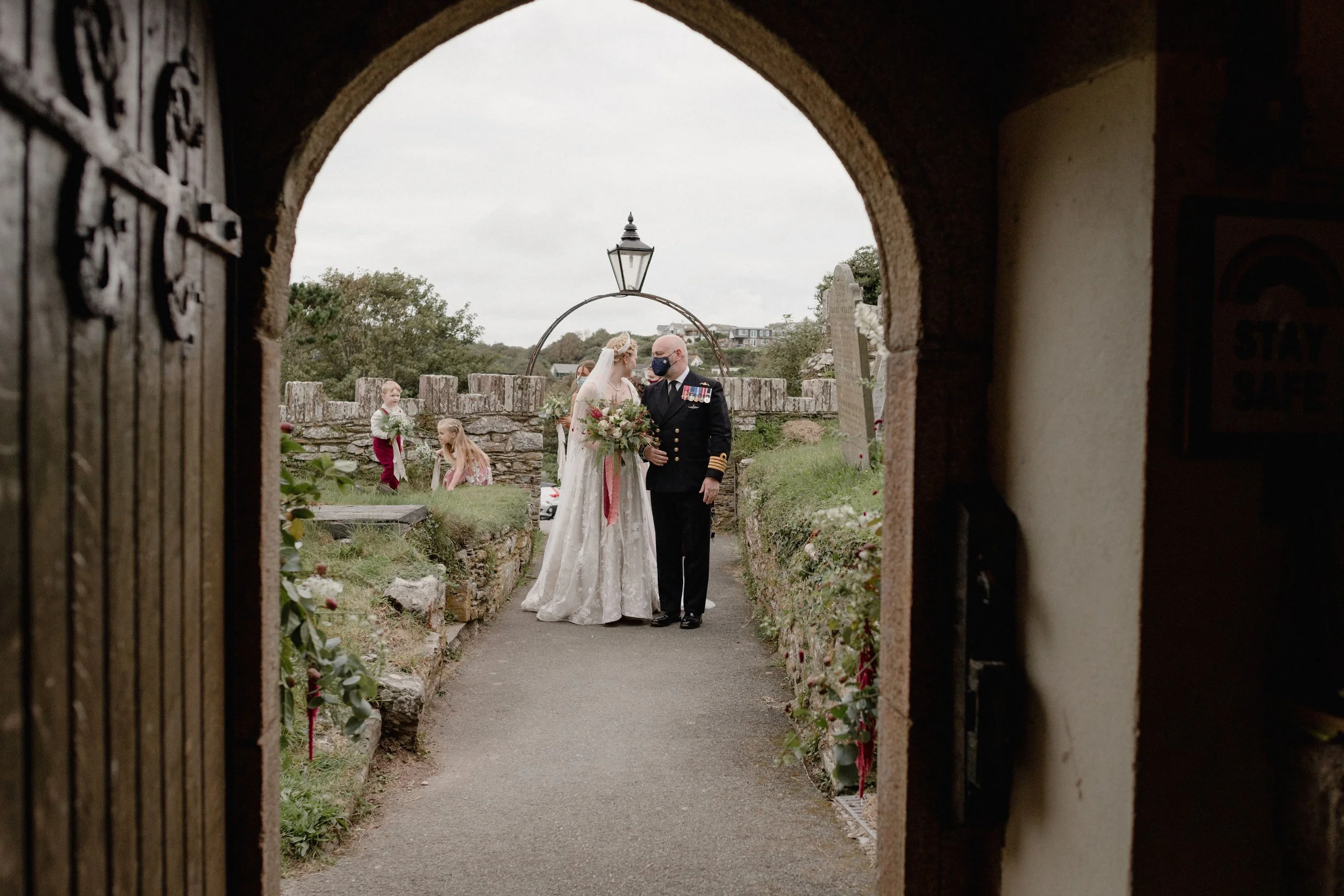 view through the church door in artistic painterly style of natural scene with children playing in the churchyard and bride and her father waiting for her devon wedding ceremony to start at Wembury