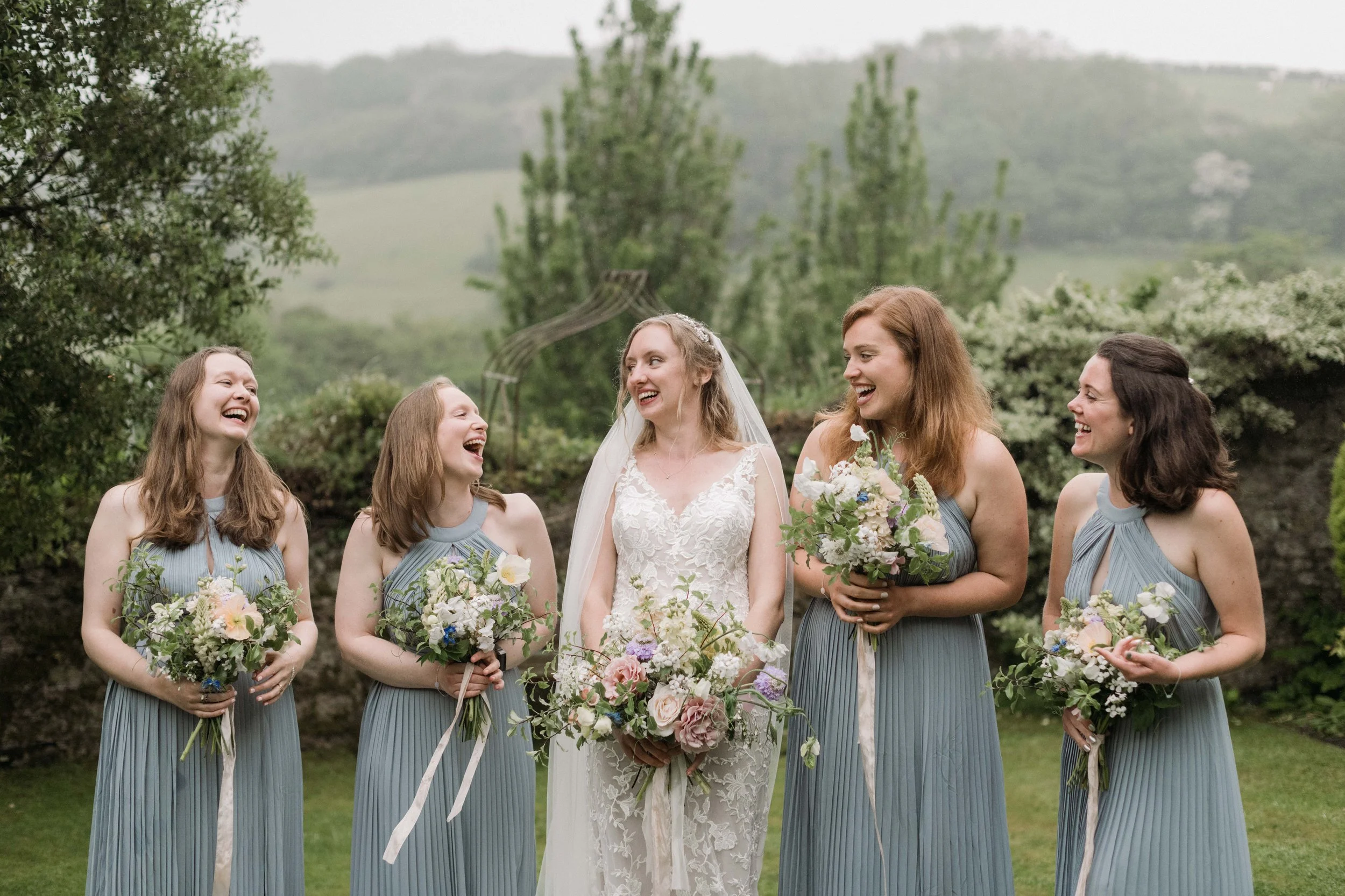 bridesmaids in the gardens with the rolling cornish hills behind them in dusky blue gowns holding bouquets by 3acre blooms