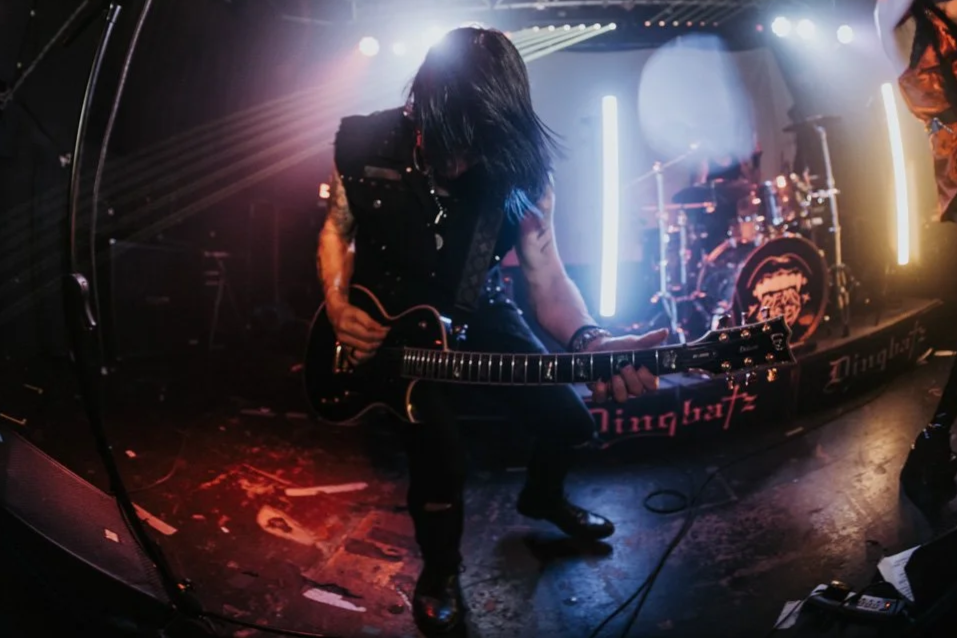 A guitarist performing on stage with a band, with lights and drums in the background.