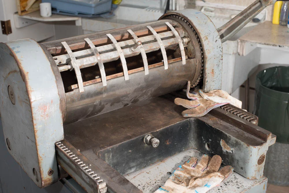 The device that spans the gap on the cylinder has a small shaft that protrudes at the middle point. This shaft has a knob on the end and that knob hits the bolt on the bed of the press right where the upper gloves pointer finger is pointing to. This acts to tip the contraption and release the paper.
