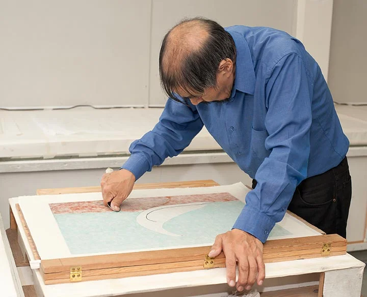 Qiatsuq spoons the paper into the ink. His hand rests on the closed registration bar.