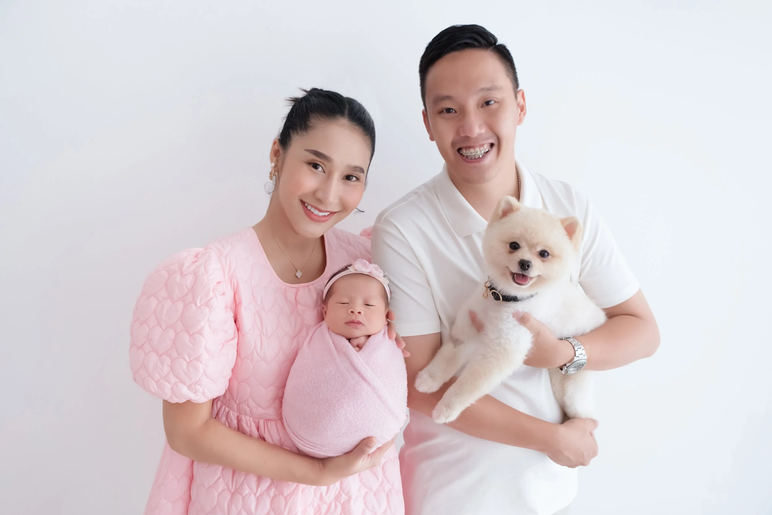 Newborn and family photography with pet by Mamamiyo Photography, Singapore: smiling parents holding their newborn and small dog, with mum wearing pink.