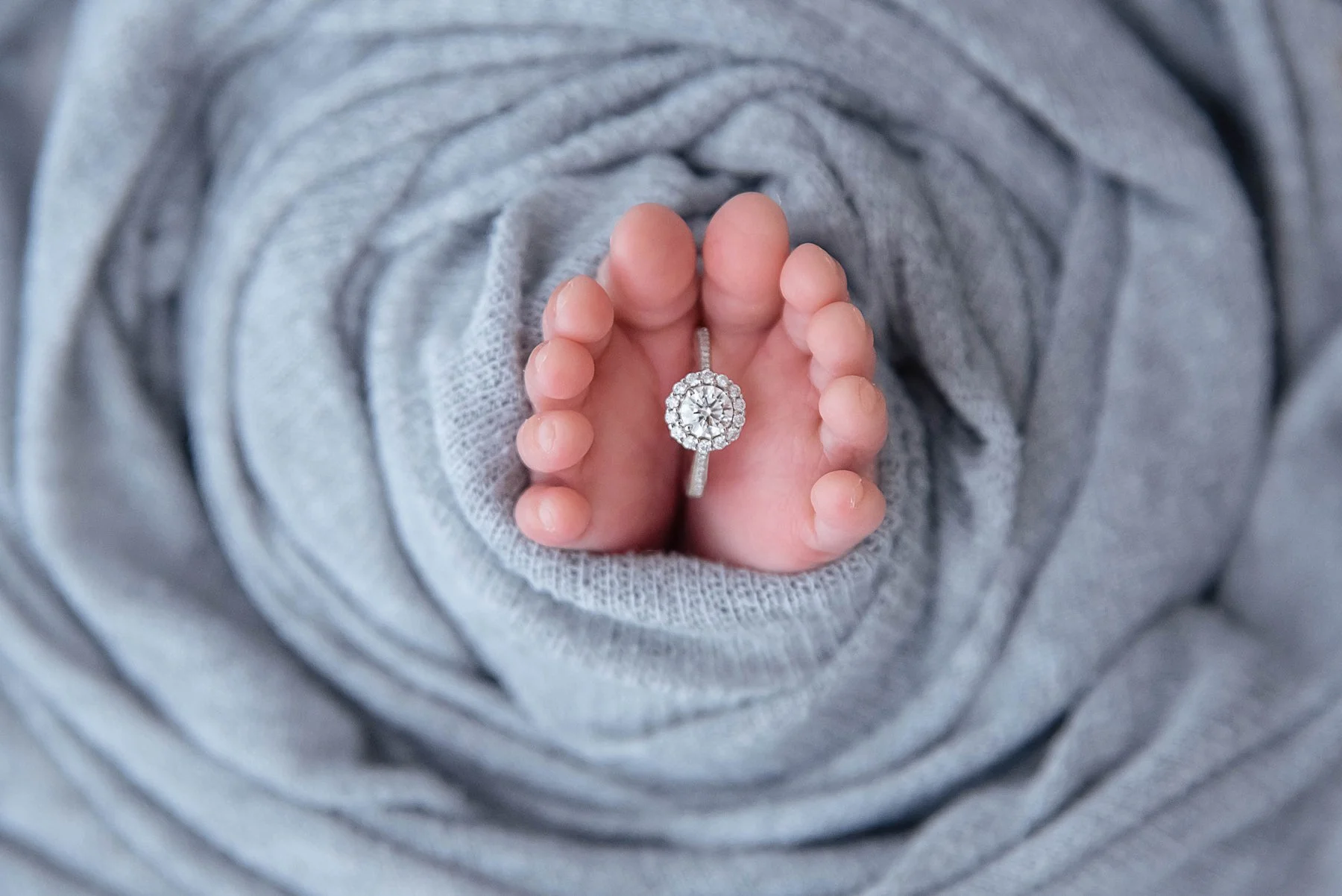 Close-up of tiny newborn feet holding a diamond ring while nestled in a grey swaddle