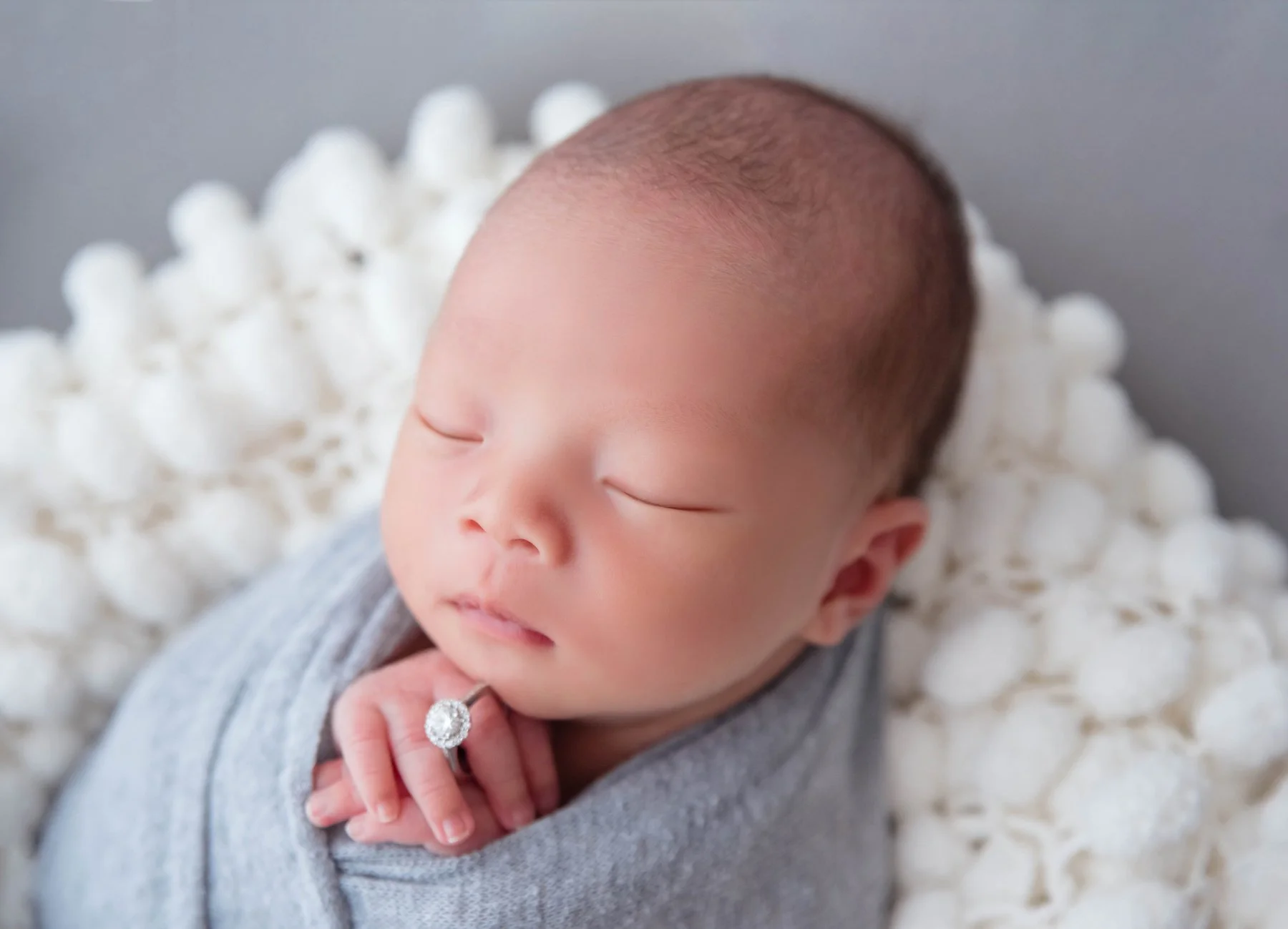 Close-up of a sleeping newborn in a grey swaddle with folded hands beneath the chin and a diamond ring resting on one finger