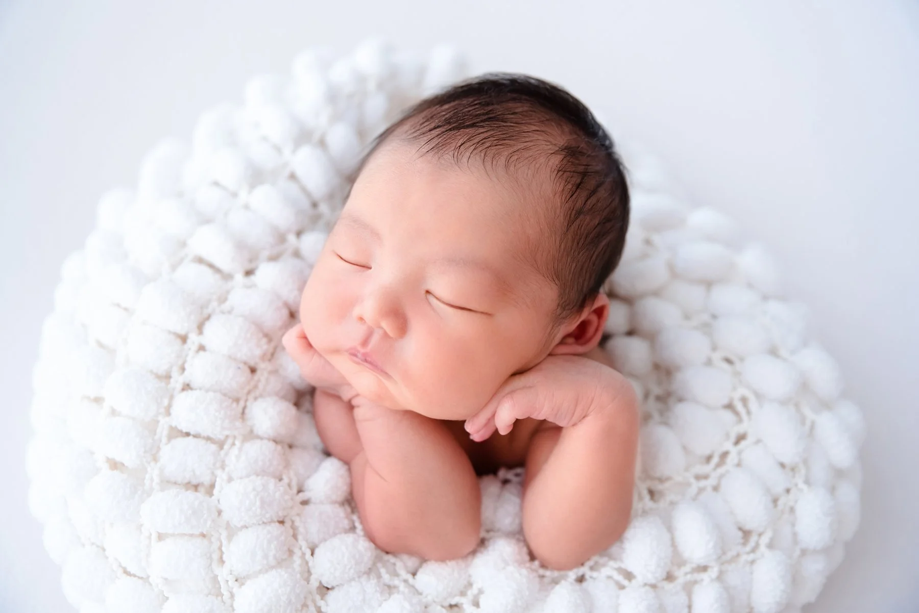 Sleeping newborn with the head turned sideways and hands beneath the chin nestled in a white knitted blanket
