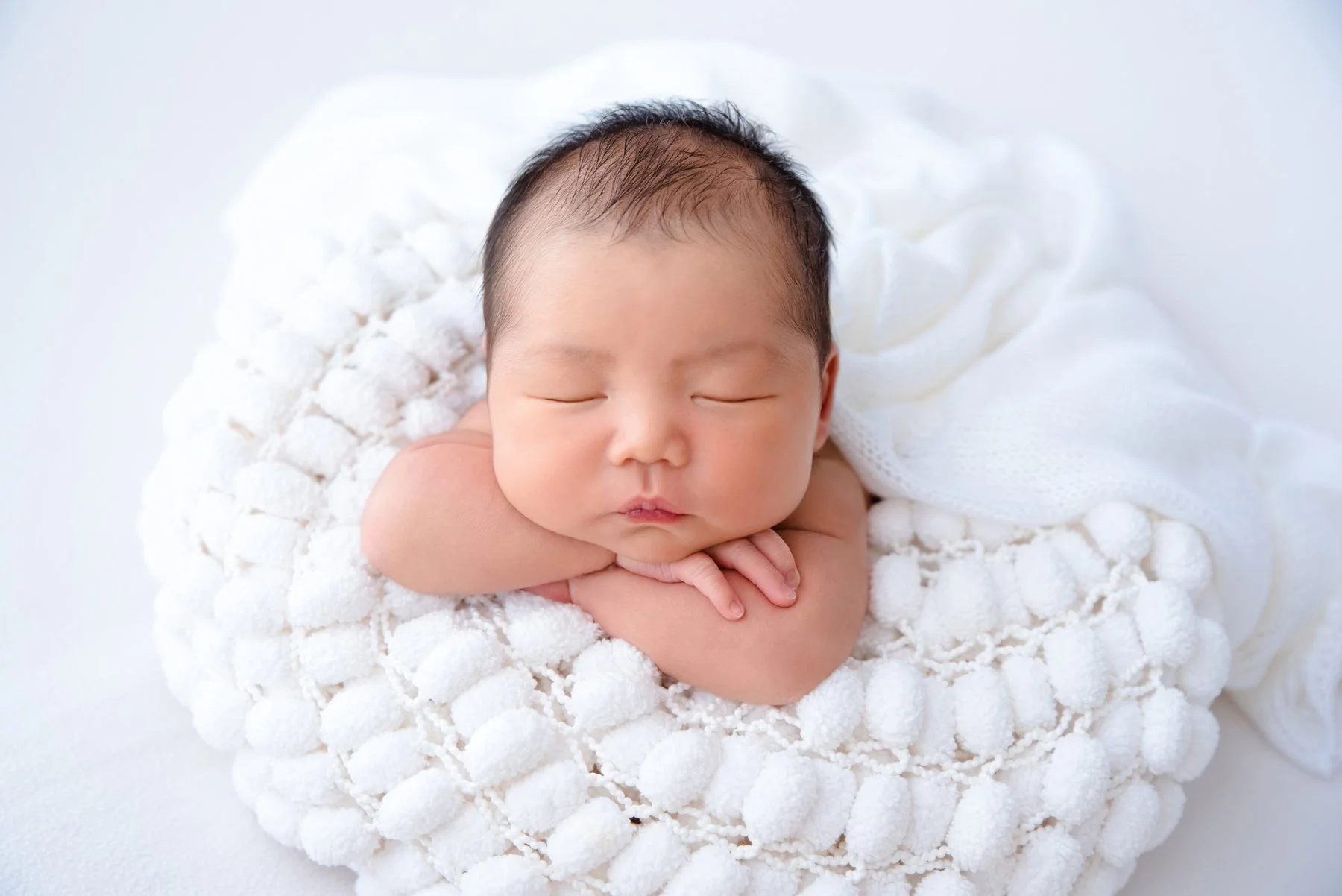 Sleeping newborn resting the chin on folded hands in a white knitted blanket against a soft white backdrop