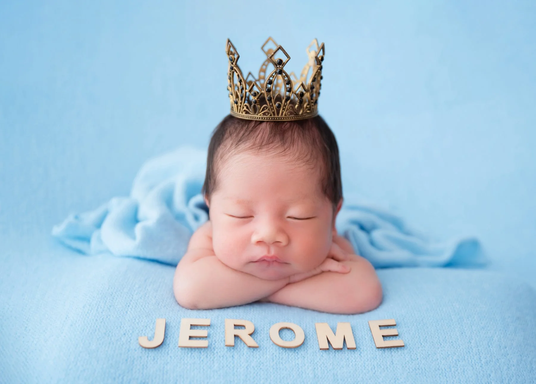 Sleeping newborn wearing a miniature gold crown resting the chin on folded arms on a blue blanket