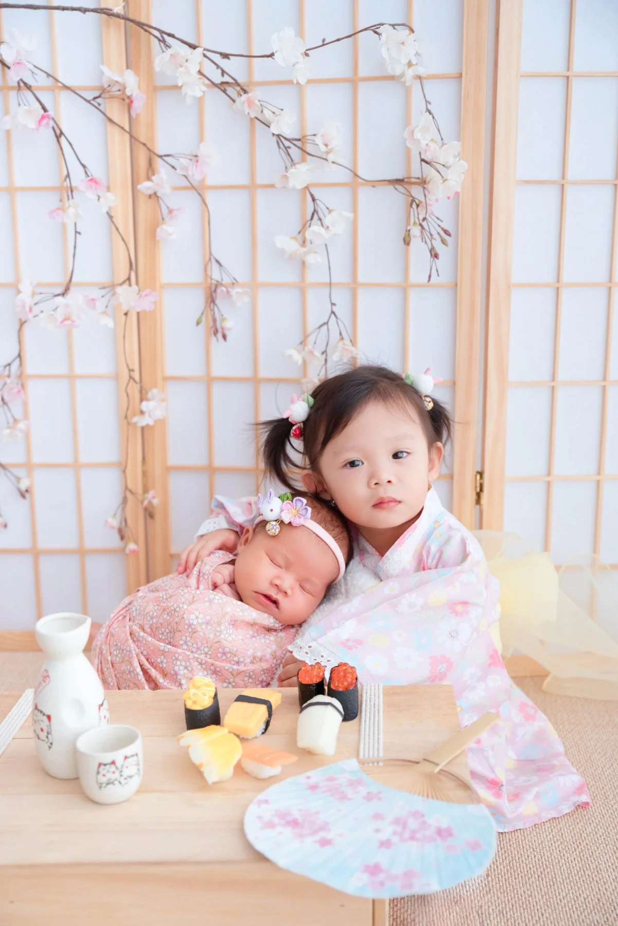 Older sister in a pink kimono holding her newborn sibling beside a Japanese-style screen