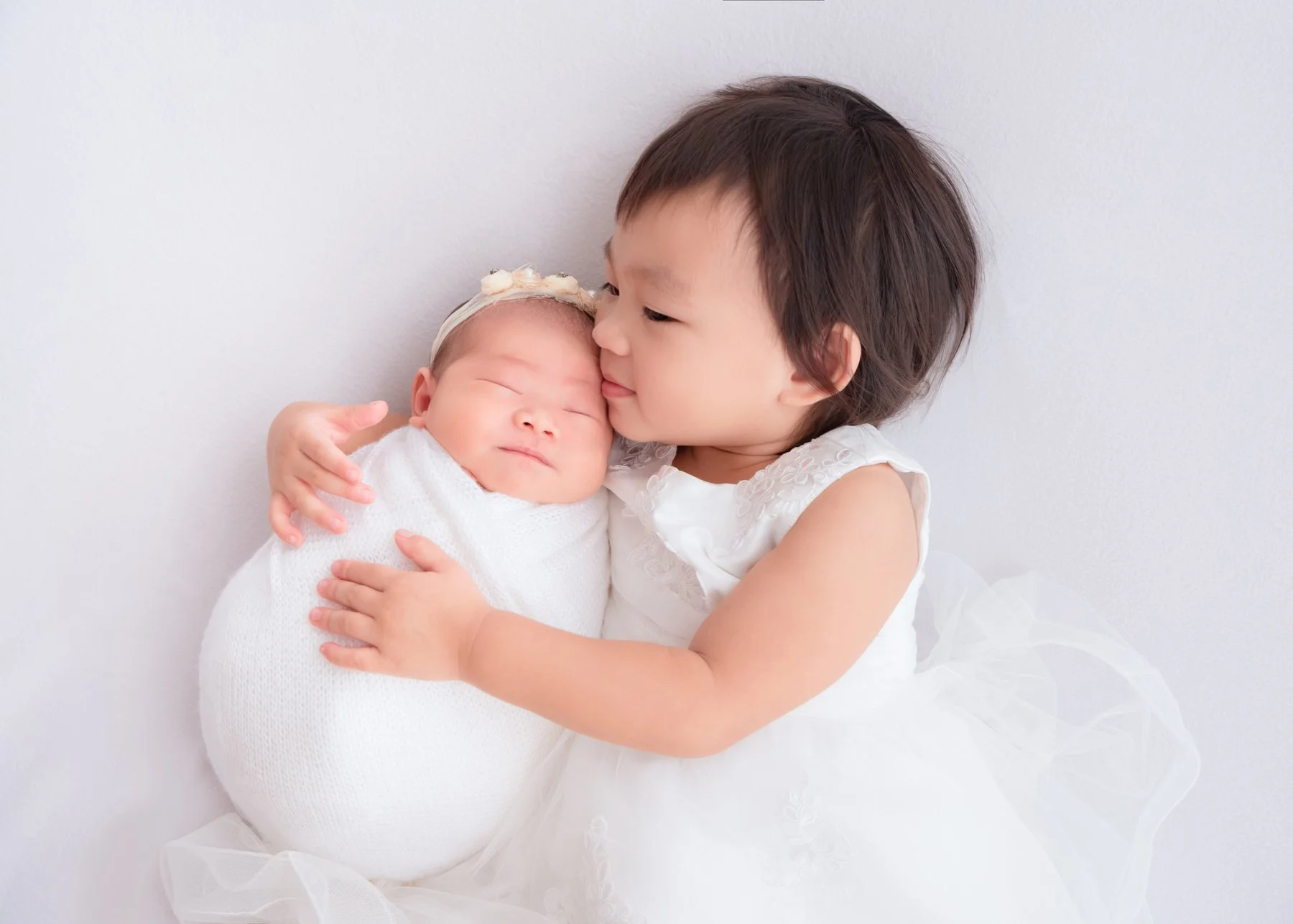 Older sister in white kissing her sleeping newborn sibling wrapped in white