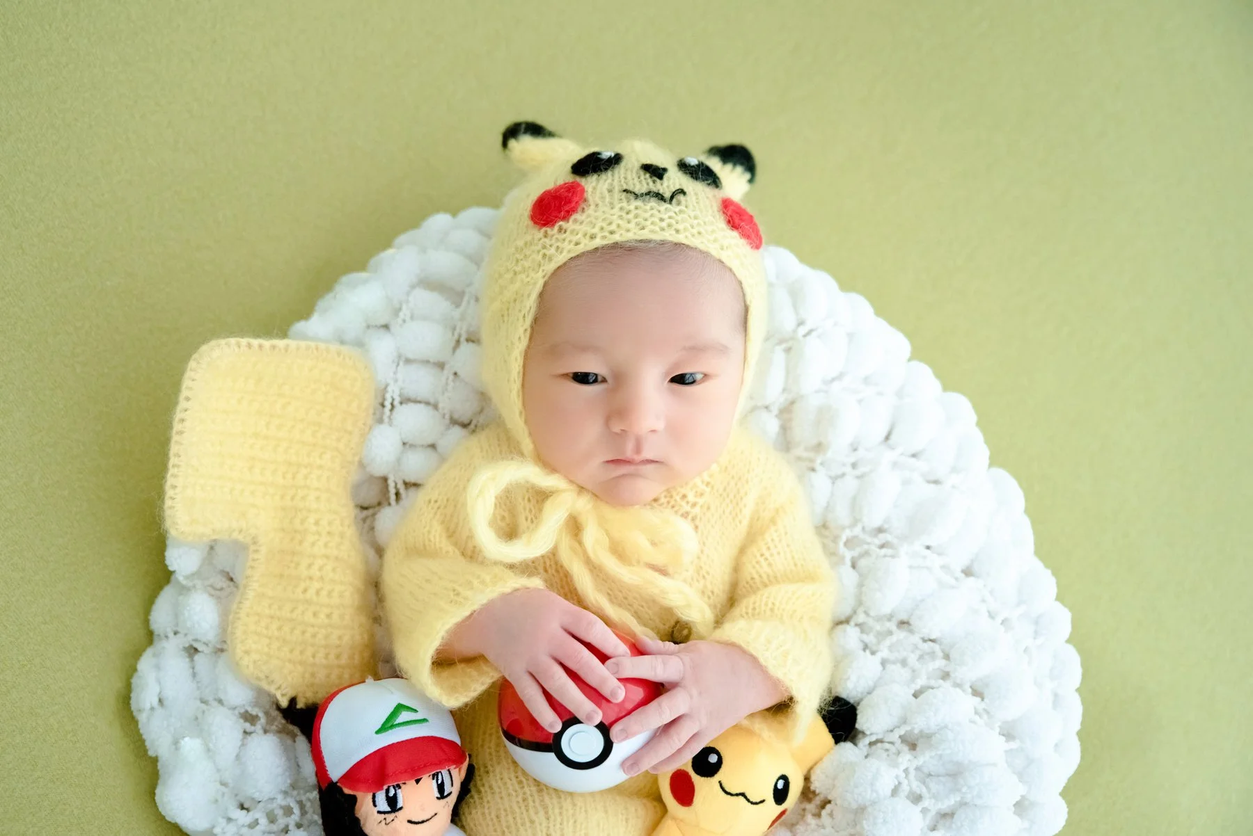 Close-up of an awake baby in a Pikachu hat and yellow outfit in a white basket with Pokémon toys