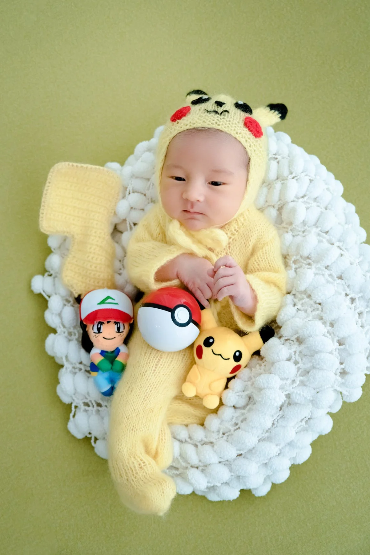 Awake baby in a yellow Pikachu outfit lying in a white basket with a Poké Ball and character toys