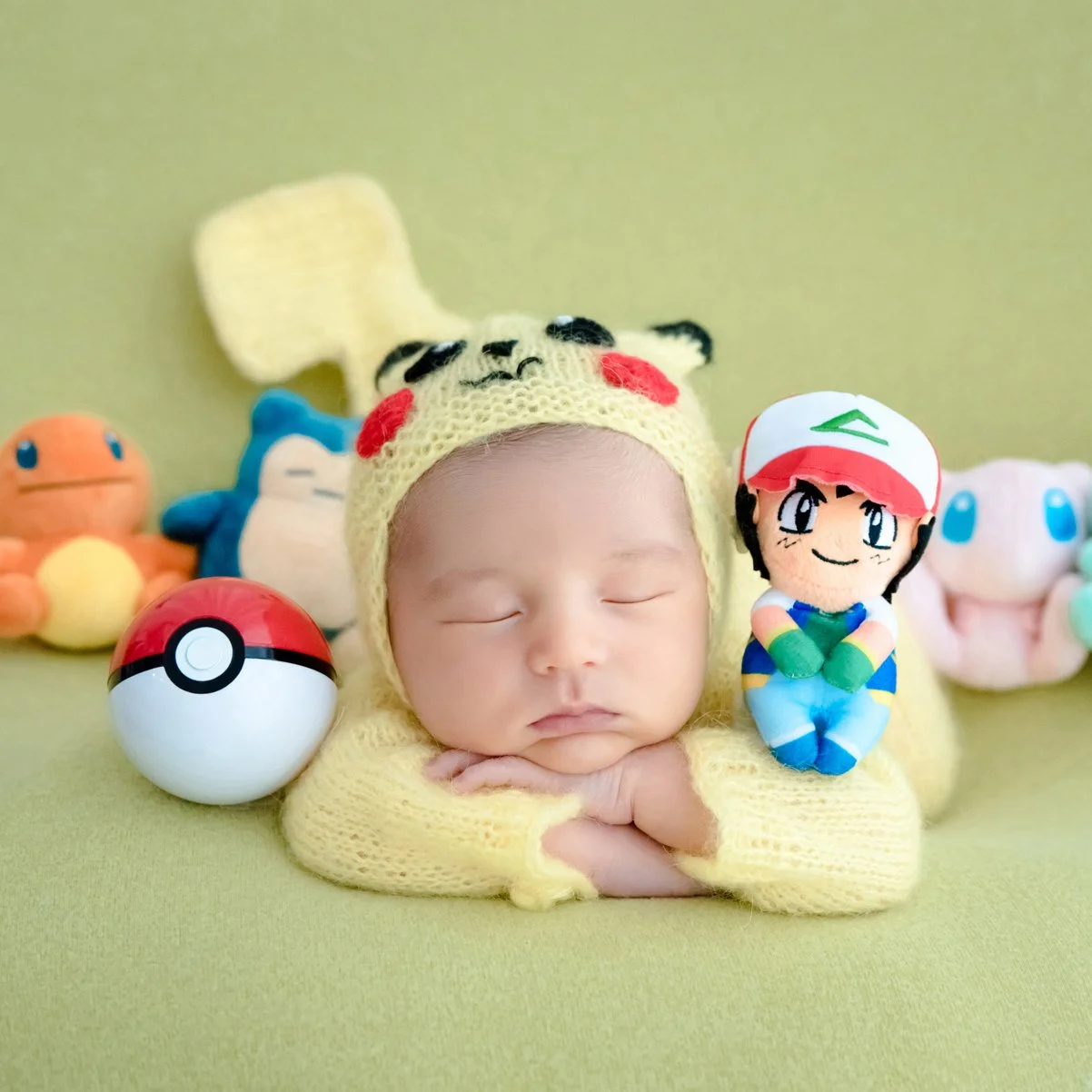 Sleeping newborn in a yellow Pikachu outfit resting the chin on folded arms with Pokémon toys behind