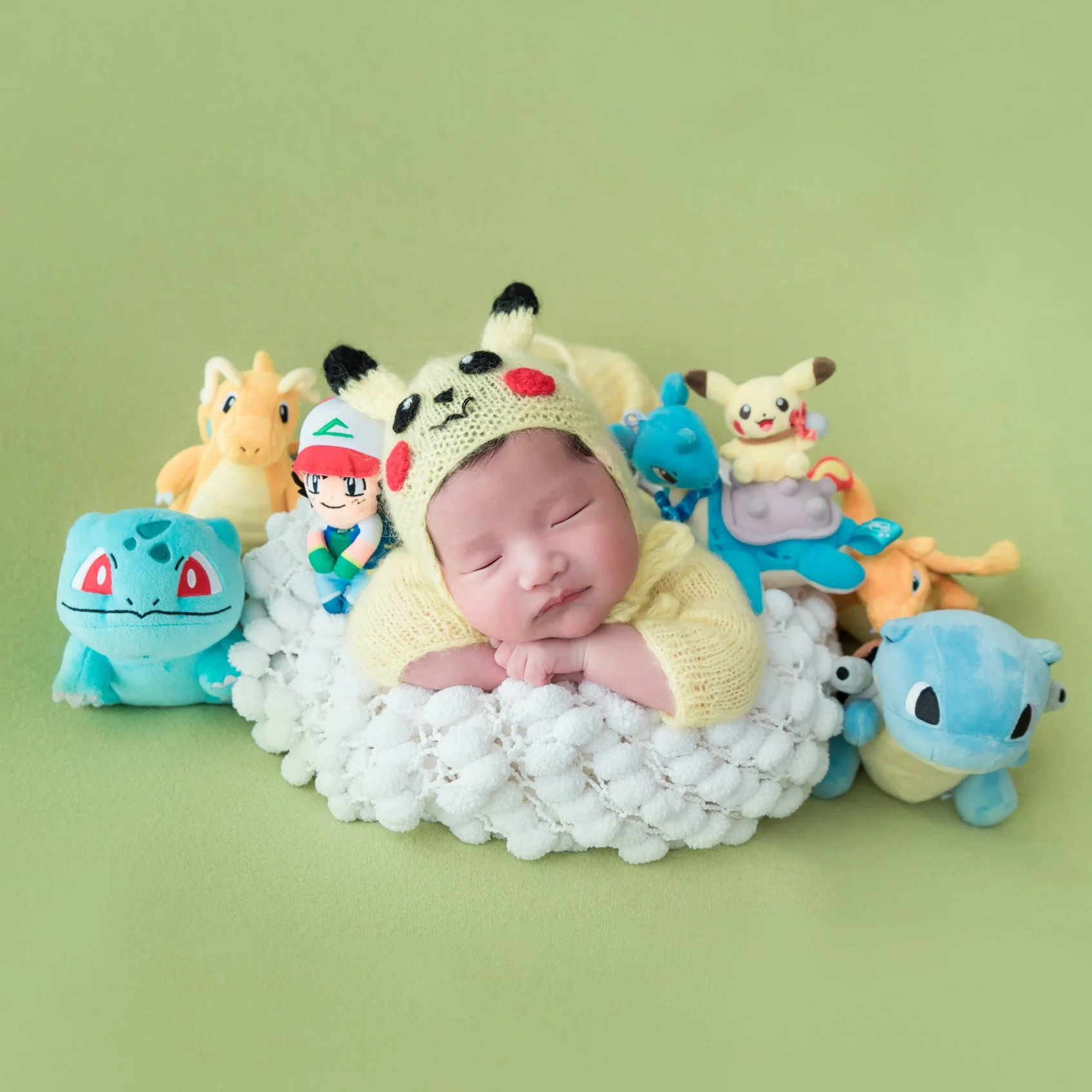 Sleeping newborn in a Pikachu hat resting in a fluffy white basket surrounded by Pokémon plush toys on green