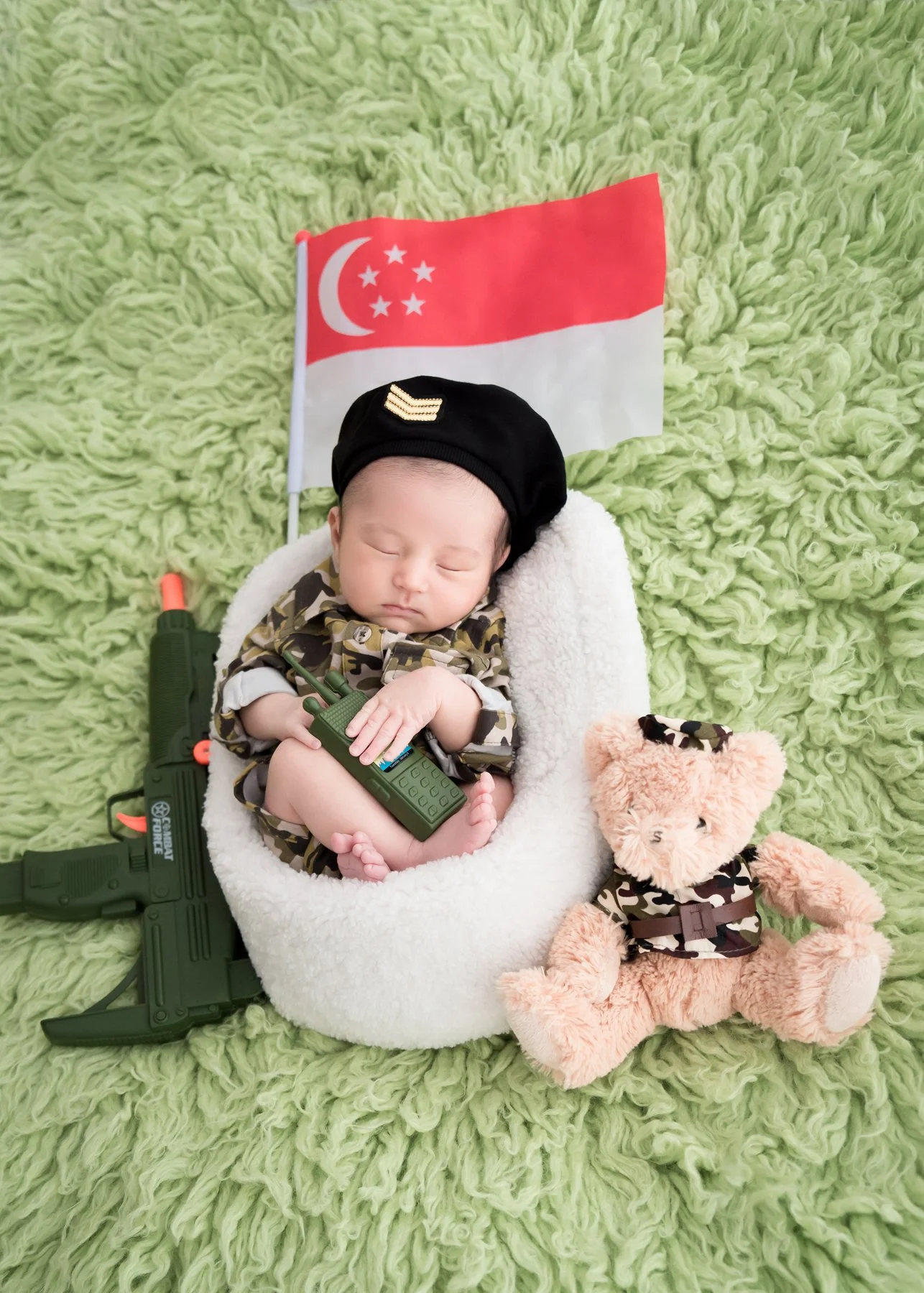 Sleeping newborn in a camouflage outfit and black beret in a white basket beside a Singapore flag, teddy bear and toy military props