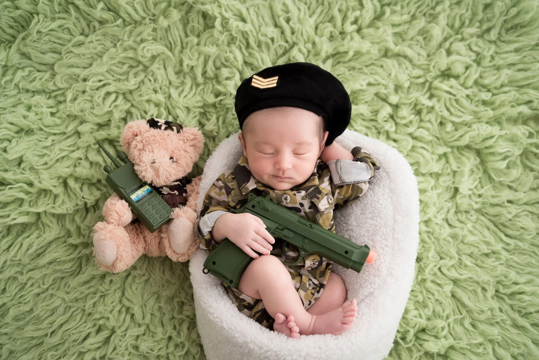 Sleeping newborn in a camouflage outfit and black beret holding a green toy rifle in a white basket beside a teddy bear