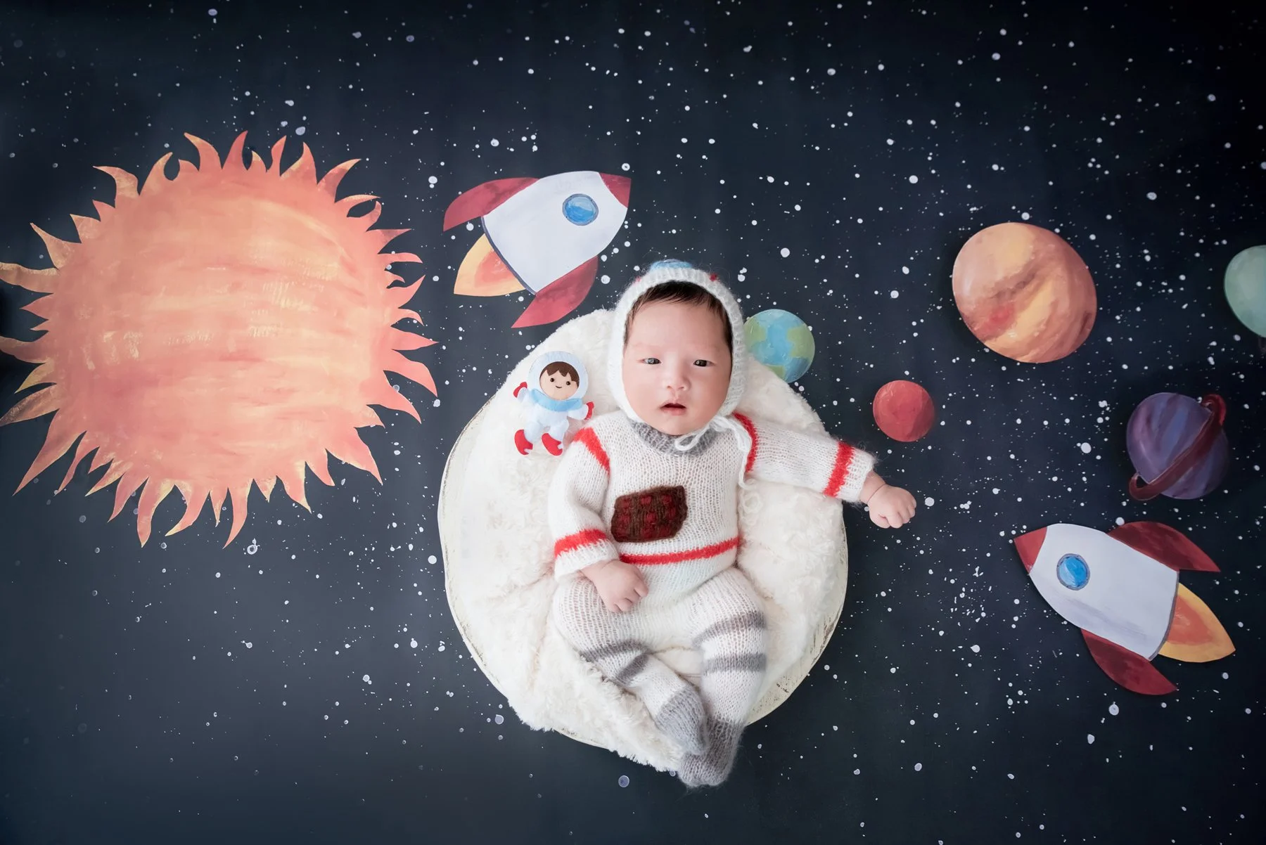 Awake newborn in a white-and-red astronaut outfit lying in a fluffy white basket on a starry backdrop with planets and rockets