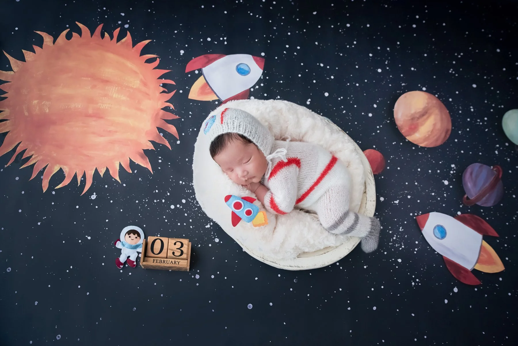 Sleeping newborn in a white-and-red astronaut outfit curled on one side in a fluffy white basket beside a toy rocket on a starry backdrop
