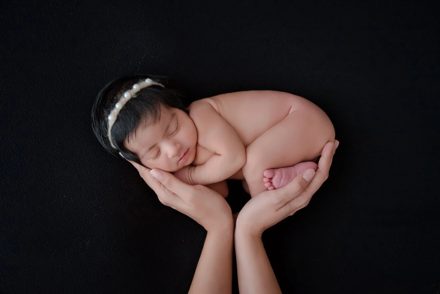 Sleeping newborn wearing a pearl headband curled on one side in adult hands against a black backdrop