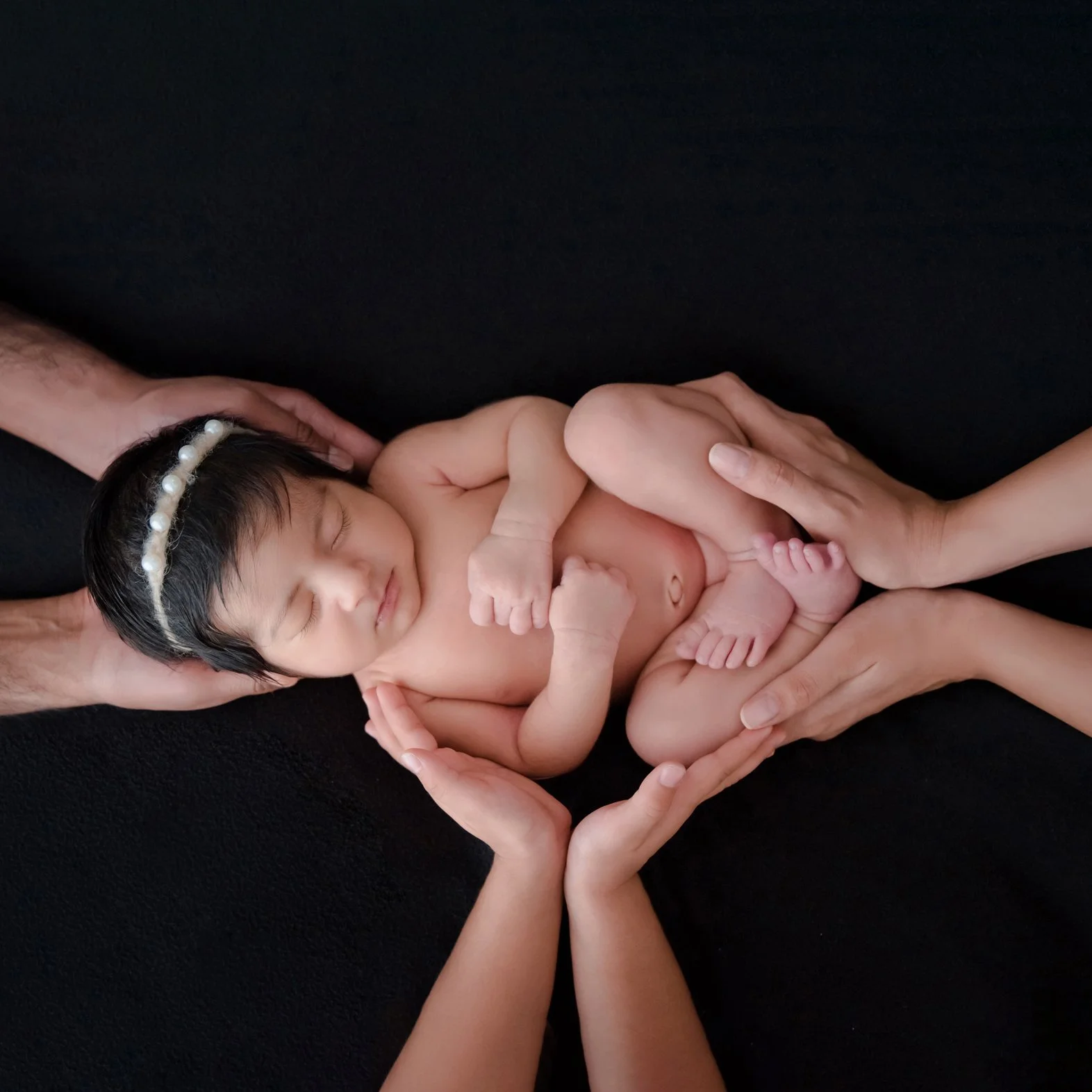 Sleeping newborn wearing a pearl headband cradled by several adult hands against a black backdrop