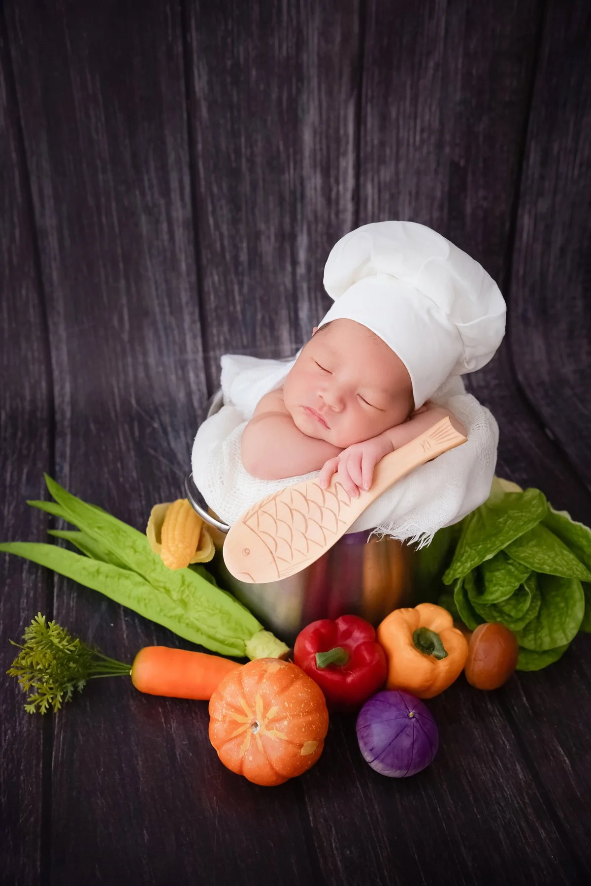 Sleeping newborn wearing a white chef hat in a small cooking pot with a wooden spoon and colourful vegetable props